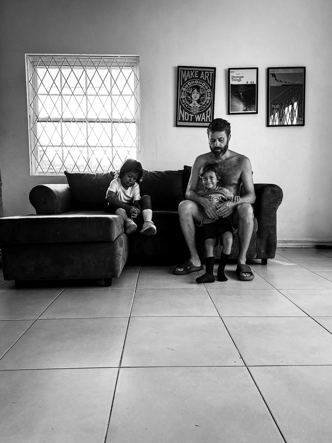 A man with a beard, two children, and a couch in a living room. The man is shirtless, sitting on the couch with a young girl on his lap. A young boy is standing in front of them, and a girl is seated on the arm of the couch. There are framed pictures