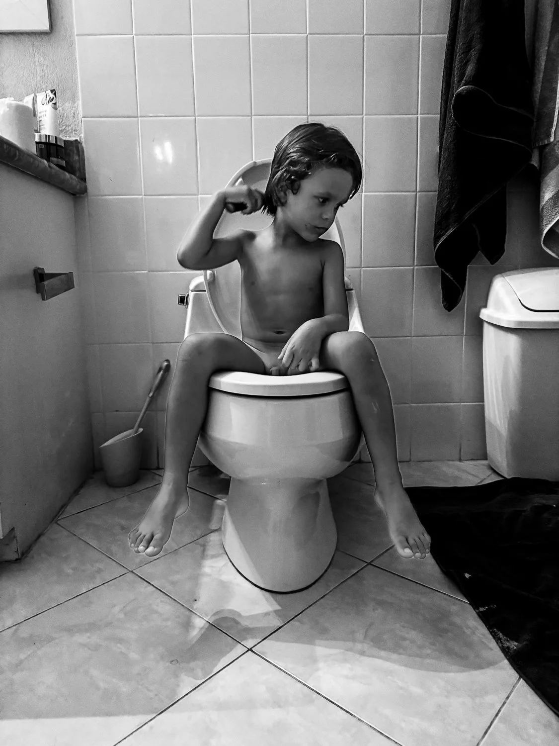 A young shirtless boy sitting on a toilet in a bathroom, holding a hairbrush in one hand, with towels hanging on the wall behind him.