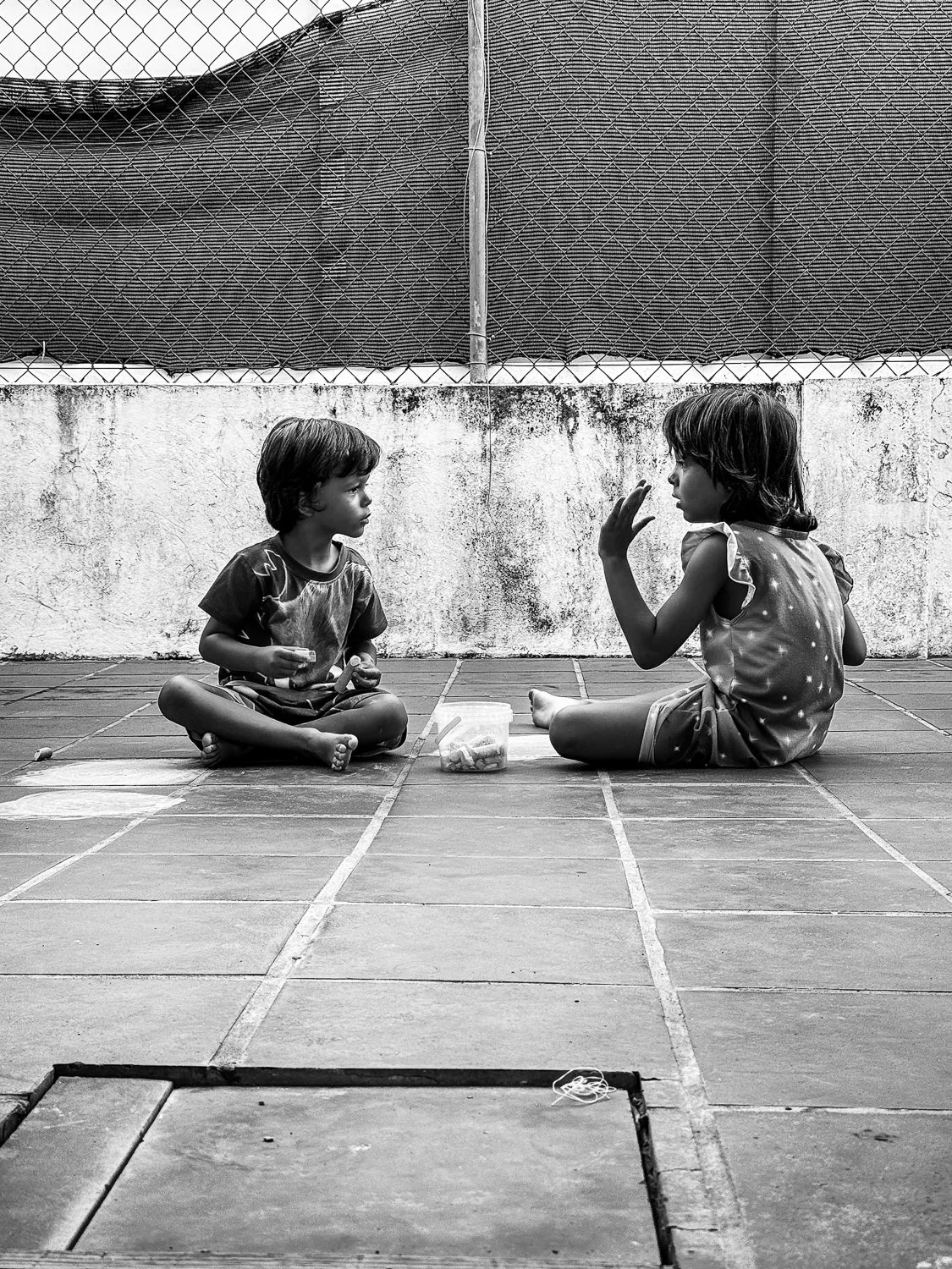 Two children sitting cross-legged on a tiled floor, facing each other, with a small container of snacks between them, and a textured wall and chain-link fence in the background.