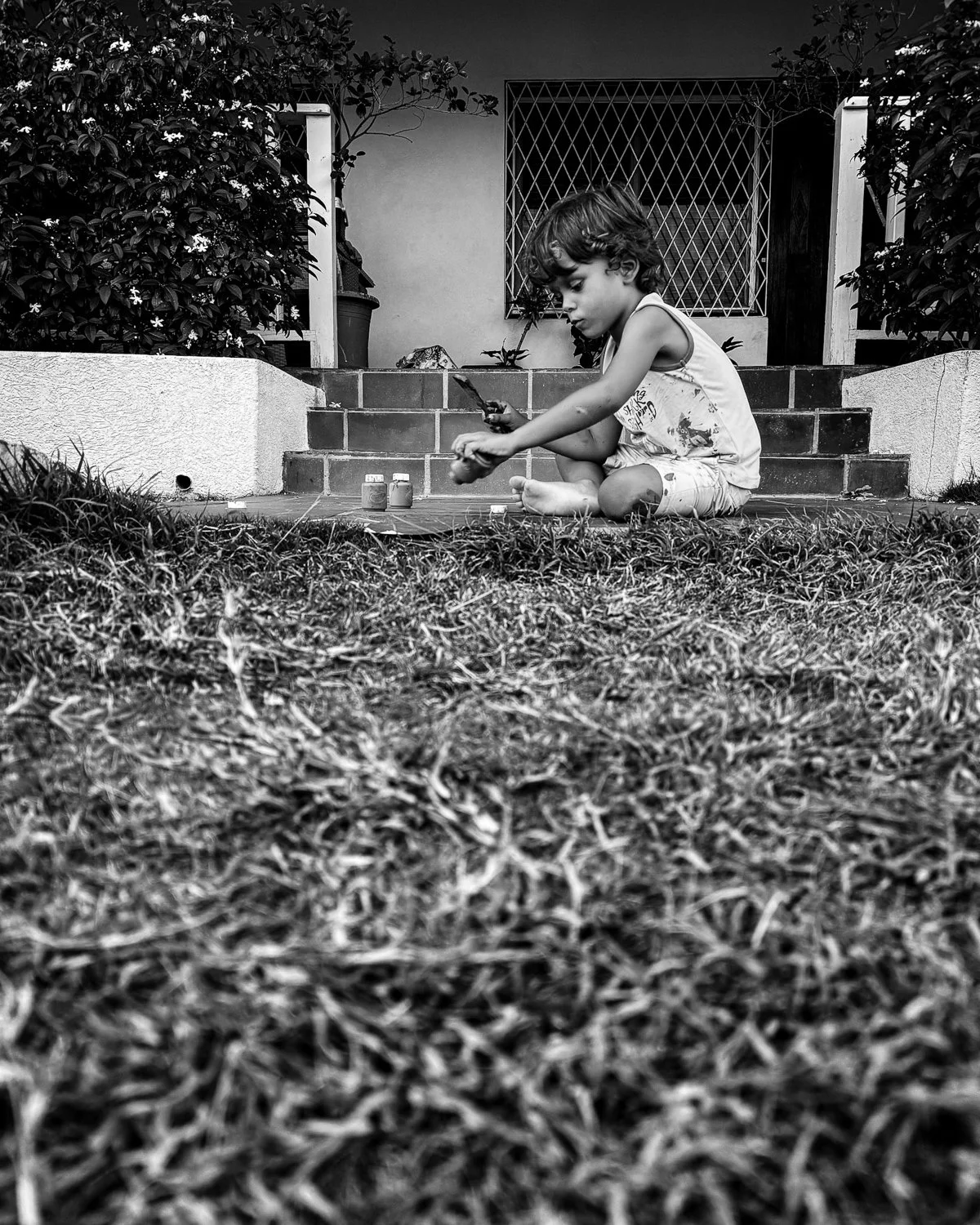 A young boy sitting cross-legged on a porch, painting or drawing on a piece of paper with small jars nearby. The porch has steps leading up to it, with plants and a window in the background. The image is in black and white.