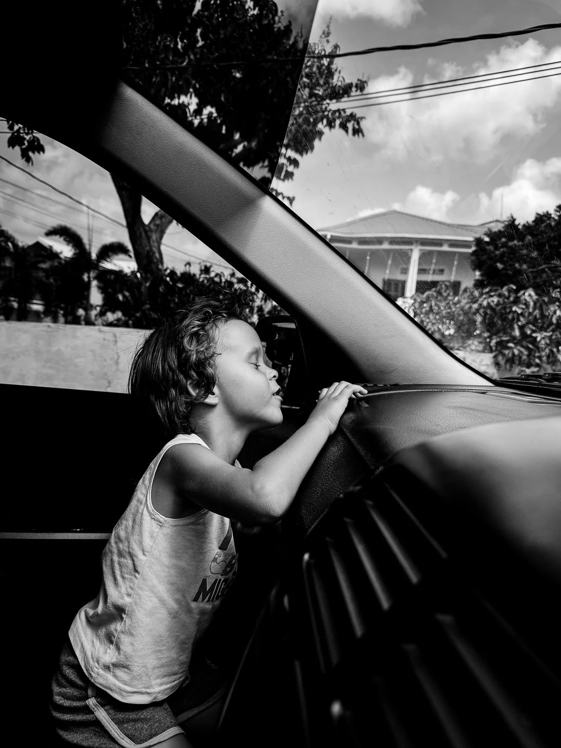A young girl with curly hair, wearing a tank top, with her eyes closed, is resting her head and hand on the dashboard of a car. The scene is captured in black and white, with a background of trees, power lines, and a house with a porch under a partly