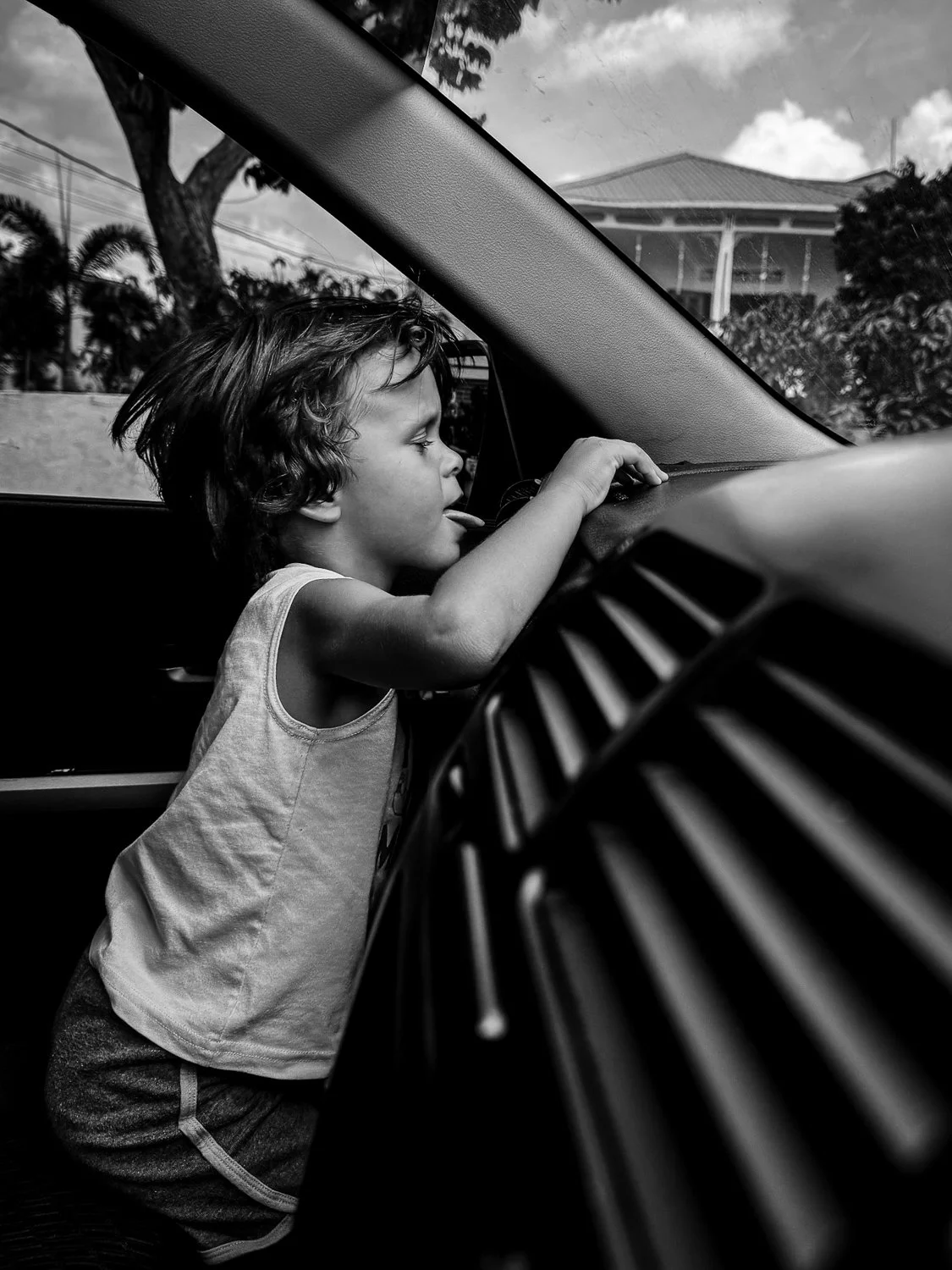 A young boy with curly hair leaning against the dashboard inside a car, looking out the front windshield with his tongue slightly sticking out. The scene is in black and white, showing trees and a house outside the car.