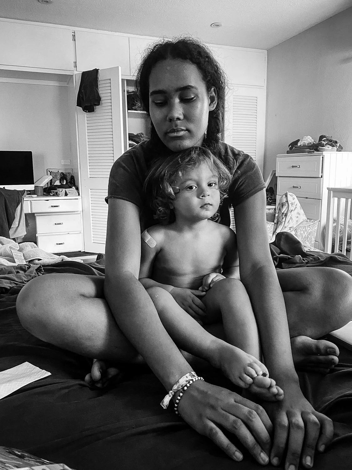 A woman sitting cross-legged on a bed holding a young child on her lap in a cluttered bedroom.