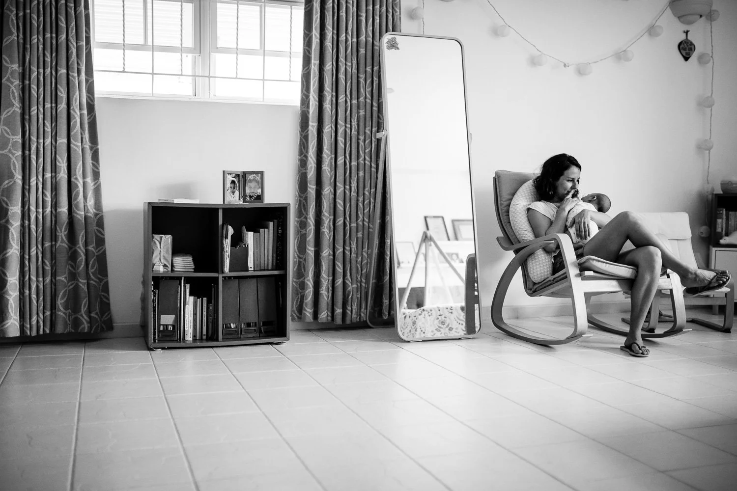 A woman sits in a rocking chair holding a baby in a bright room with a large window, curtains, a mirror, a bookshelf, and decorative string lights.