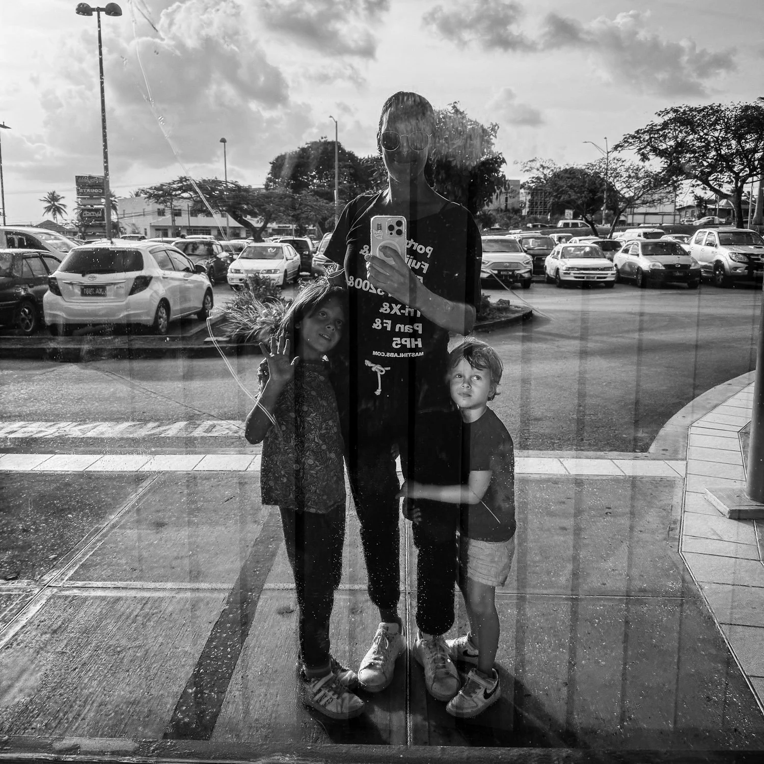 A woman with two children taking a selfie in a parking lot reflection on a glass door, with cars and trees in the background.