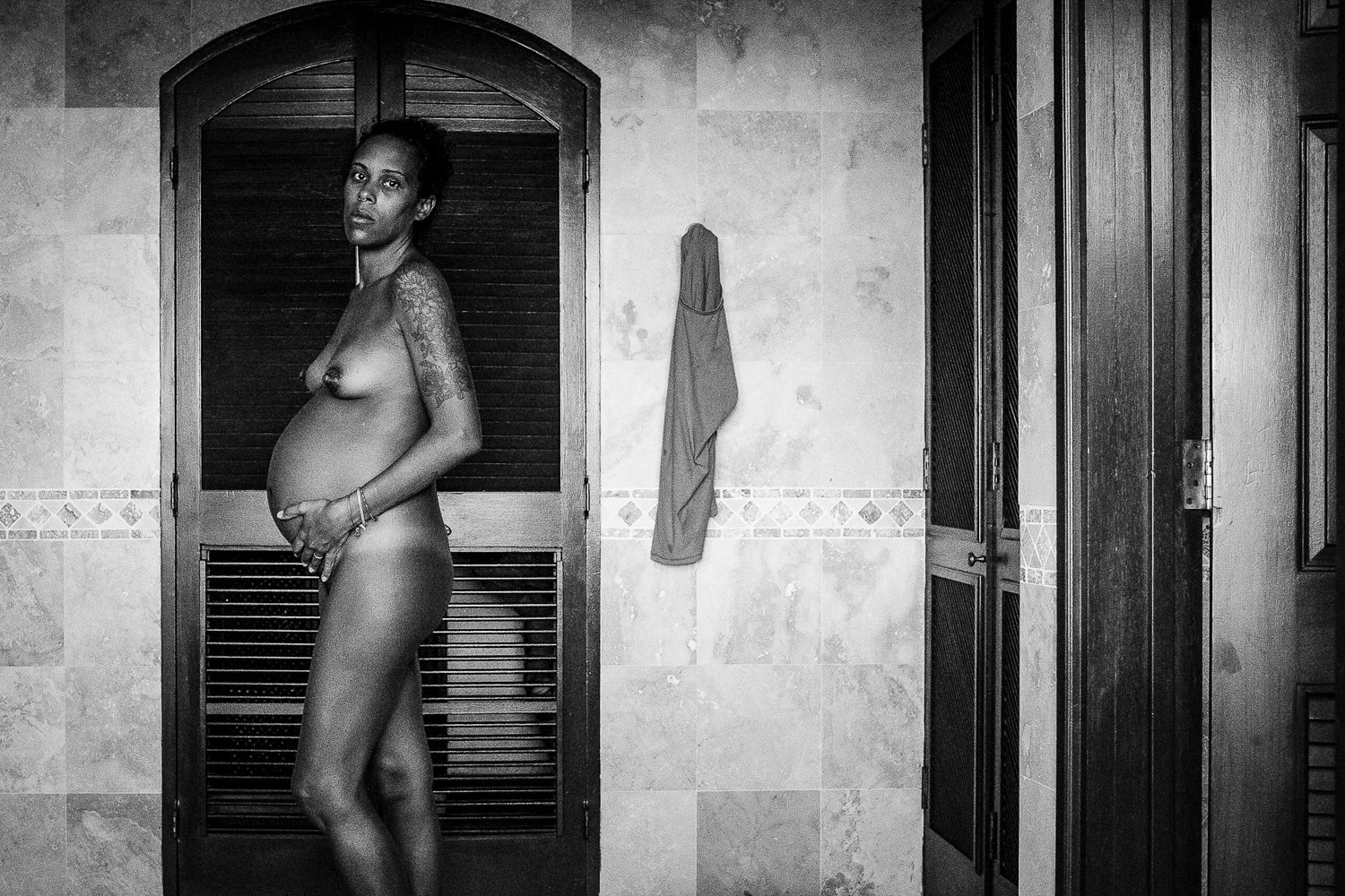 A pregnant woman standing in a bathroom, nude, with a tattooed arm, holding her belly, looking at the camera. The bathroom has tiled walls, a hanging towel, and wooden louvered doors.