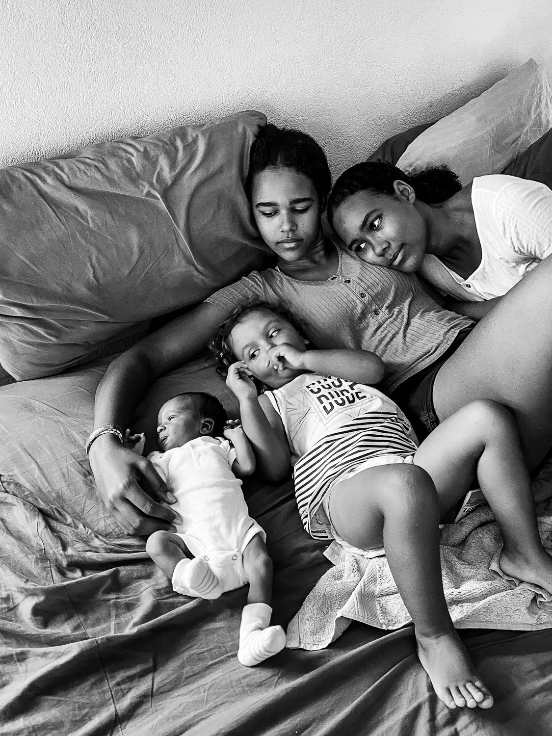 Two women and two children lying on a bed, relaxing together. The older girl and woman are resting their heads on pillows, with the girl looking thoughtfully, while the woman gazes at her. The younger girl is lying on her back, looking up, and the in