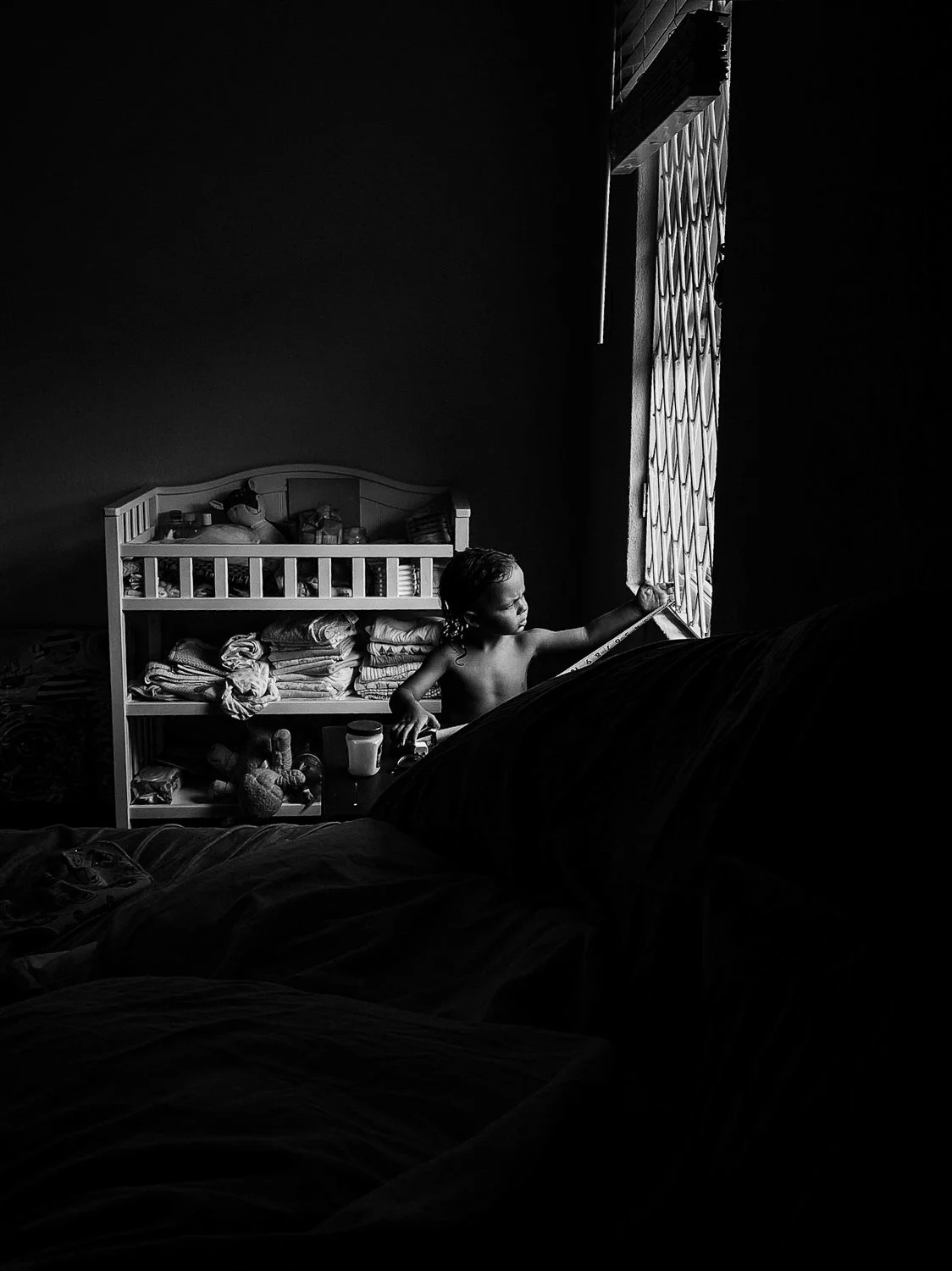 A young child looking out of a window, standing next to a bed and a shelf with folded clothes and toys, in a dimly lit room.