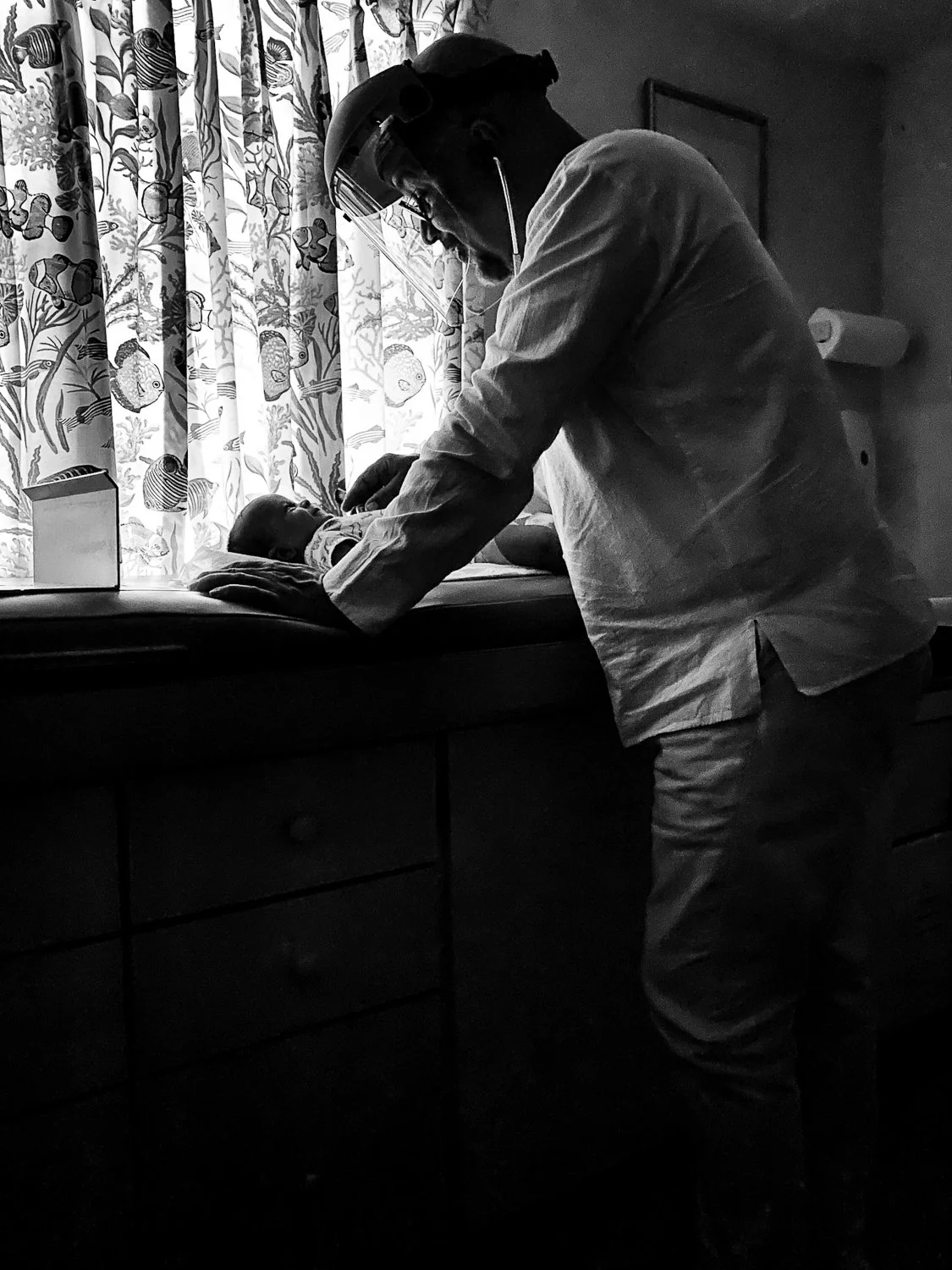 A healthcare professional in protective gear examines a newborn baby on an examination table in a hospital room with patterned curtains in the background.