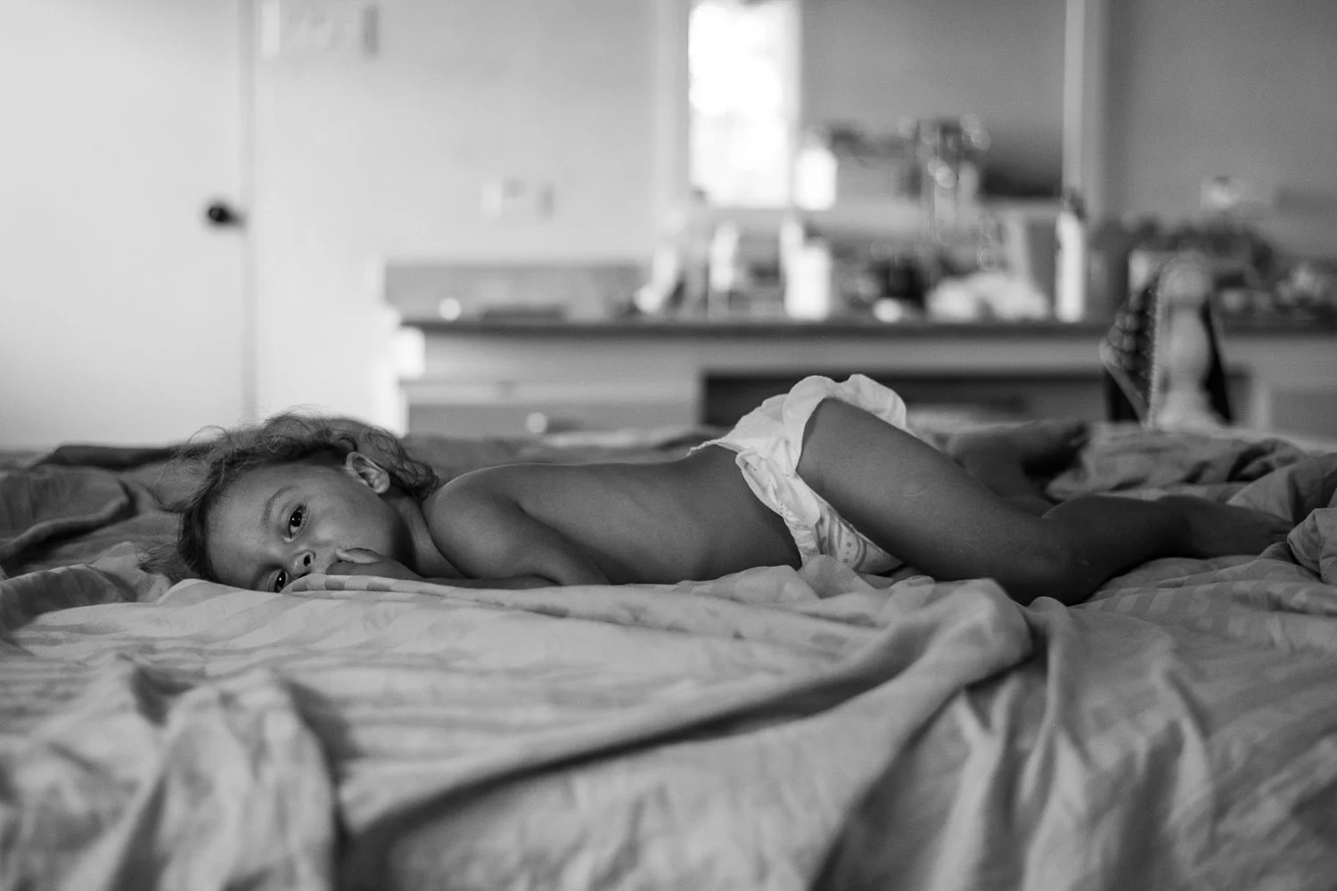 A young girl with curly hair lying on her side on a bed, looking towards the camera, wearing a diaper, in a black and white photo.