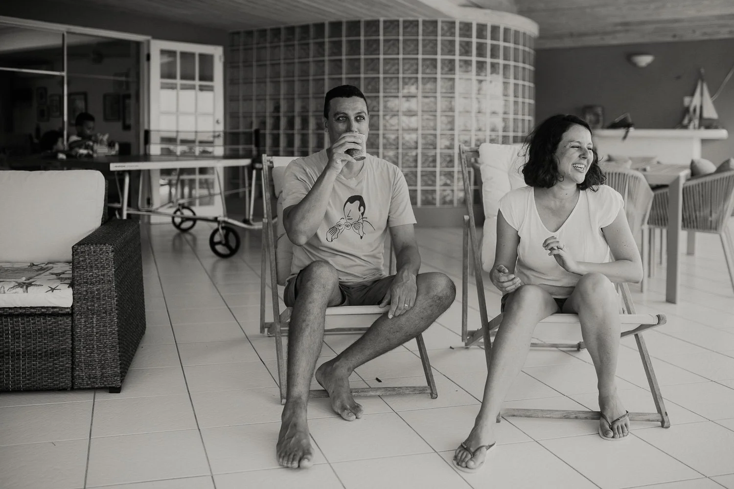 A black and white photo of a man and woman sitting in chairs playing a game of Jenga indoors. The man is drinking from a cup, wearing a t-shirt with a cartoon face, shorts, and barefoot. The woman is smiling, wearing a white t-shirt, shorts, and flip