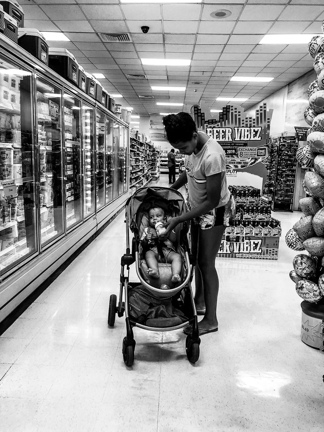A woman is pushing a stroller with a baby sitting inside in a grocery store aisle. The woman is leaning over the stroller, feeding the baby. The store has shelves stocked with products and there are promotional signs in the background.