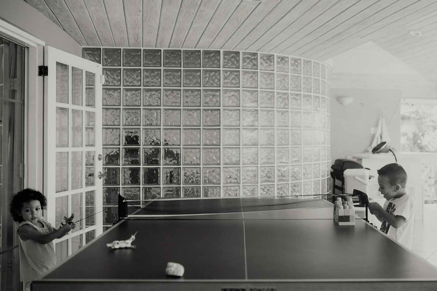 Two children playing table tennis indoors. The girl with curly hair is holding a paddle and looking at the camera, while the boy on the other side is also holding a paddle and focused on the game. The room has a glass block wall, a door with glass pa