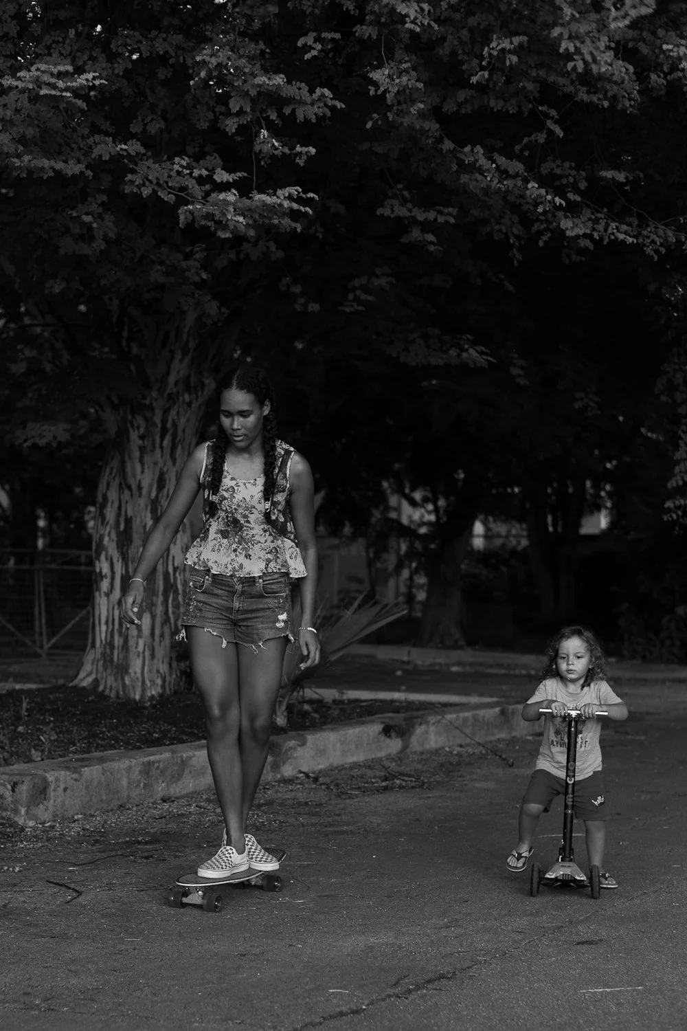 A young girl rides a scooter while an older girl skateboards on a street with large trees in the background.