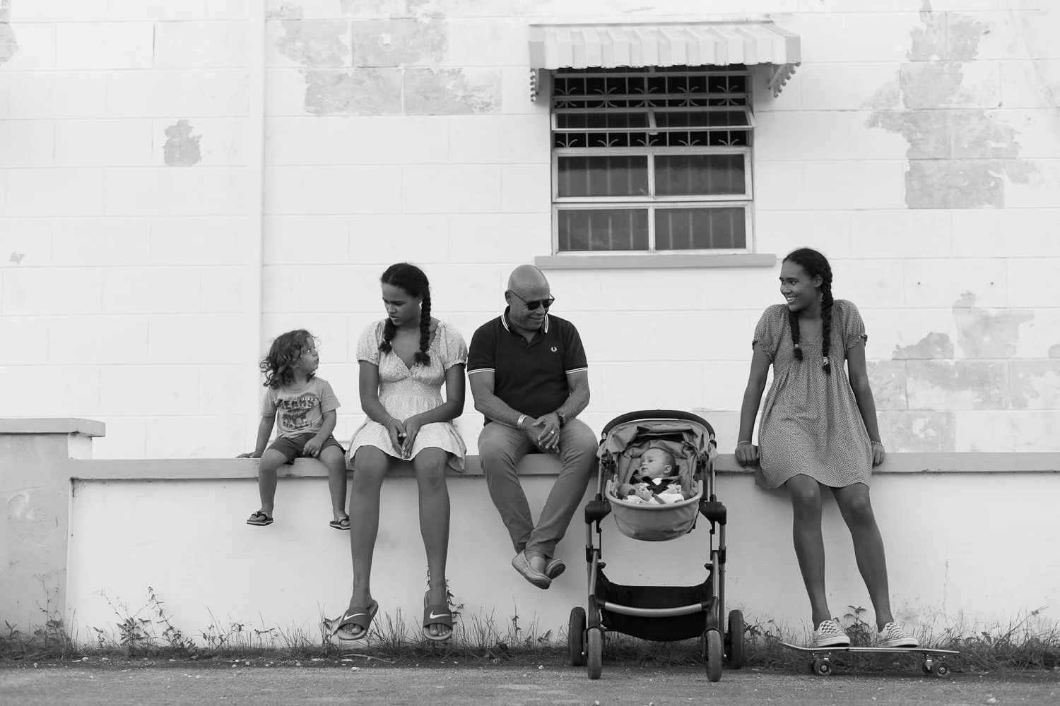 A black-and-white photo of five family members sitting on a low wall in front of a building with a barred window. From left to right: a young girl sitting on the wall, a woman sitting with her hands clasped, a man wearing sunglasses and sitting with
