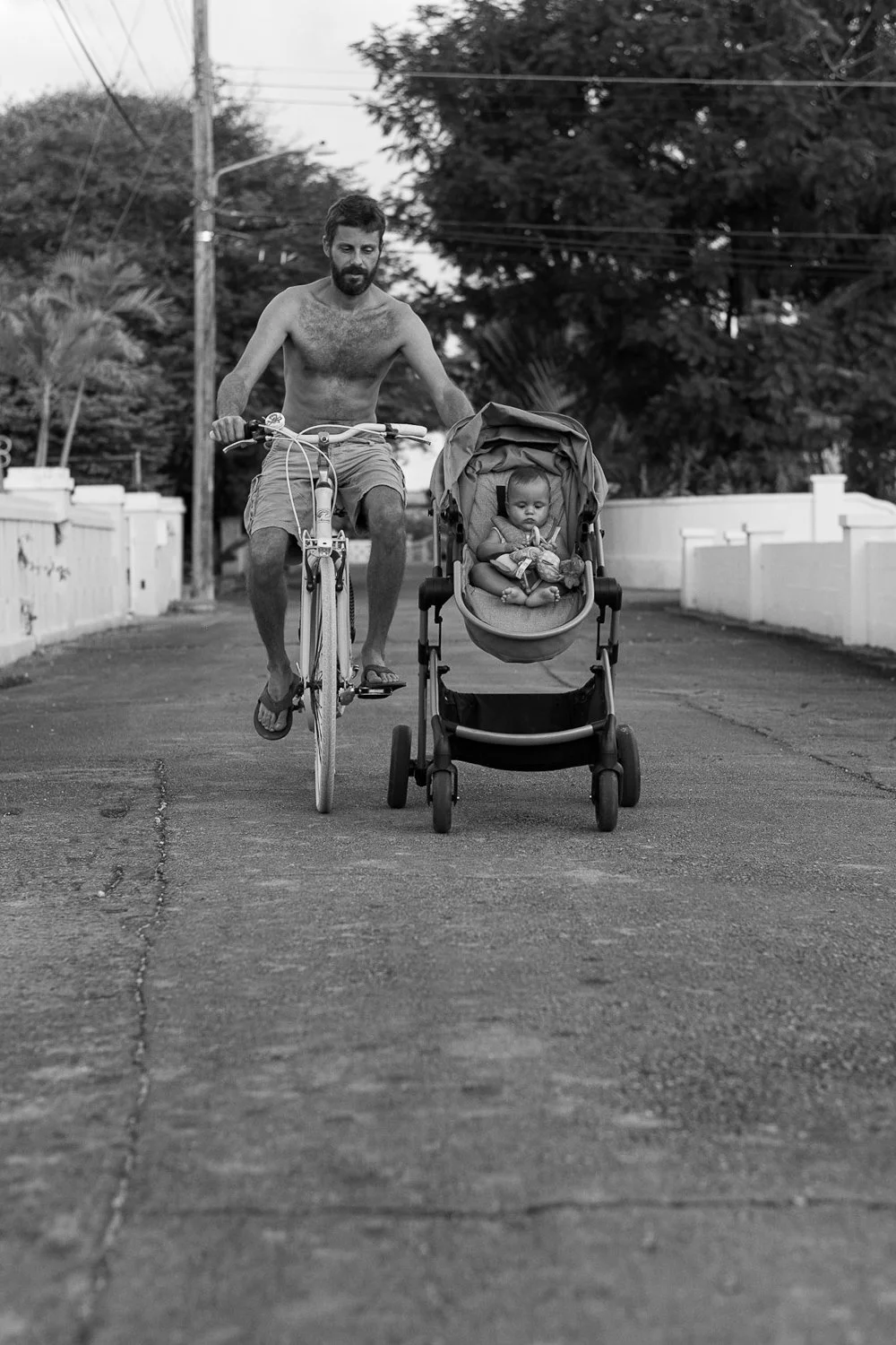 A man riding a bicycle next to a baby stroller on a street, with a young child seated in the stroller.
