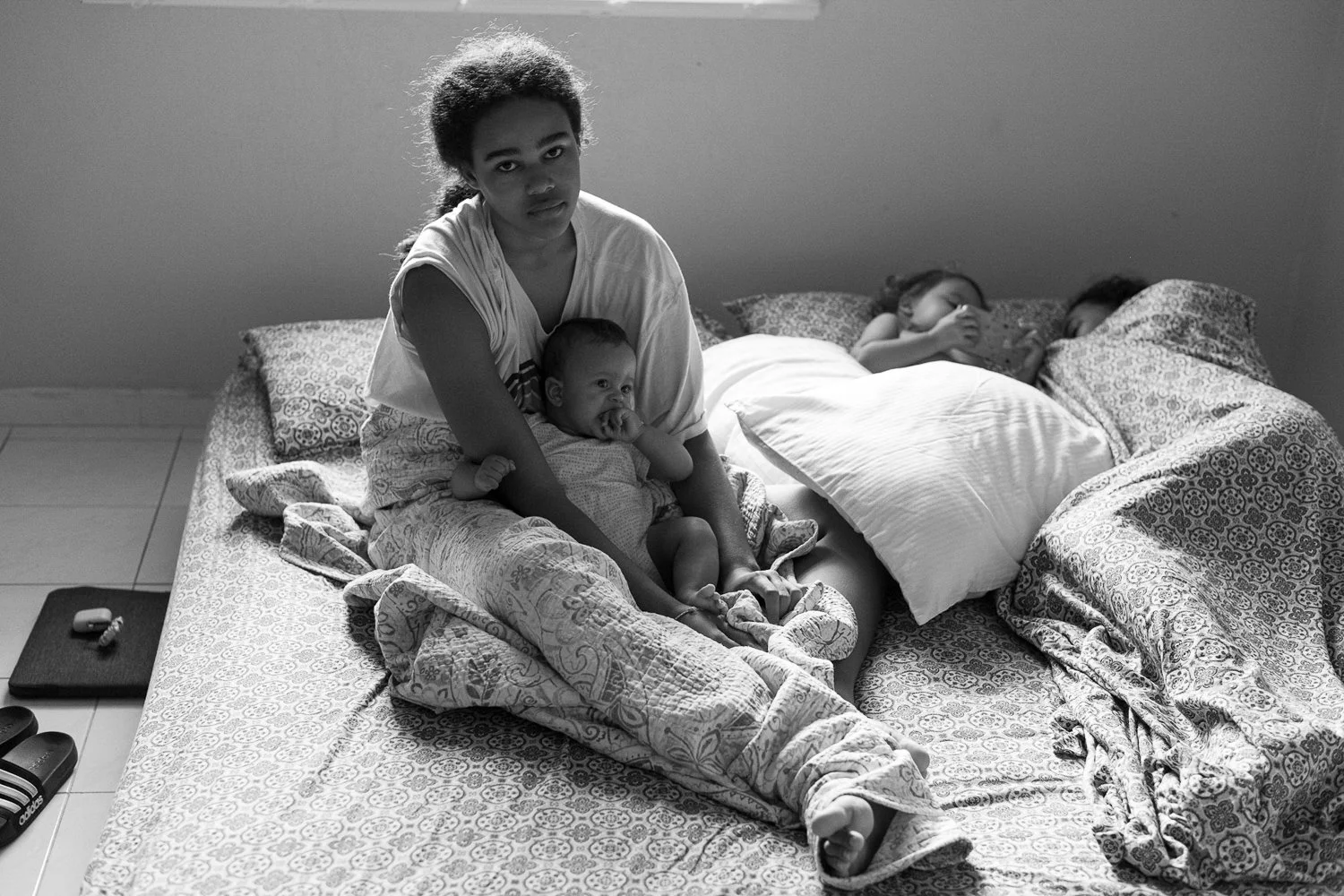 A woman sits on a bed with three children, one of whom is a baby, while two others are sleeping in bed. The scene is in black and white.