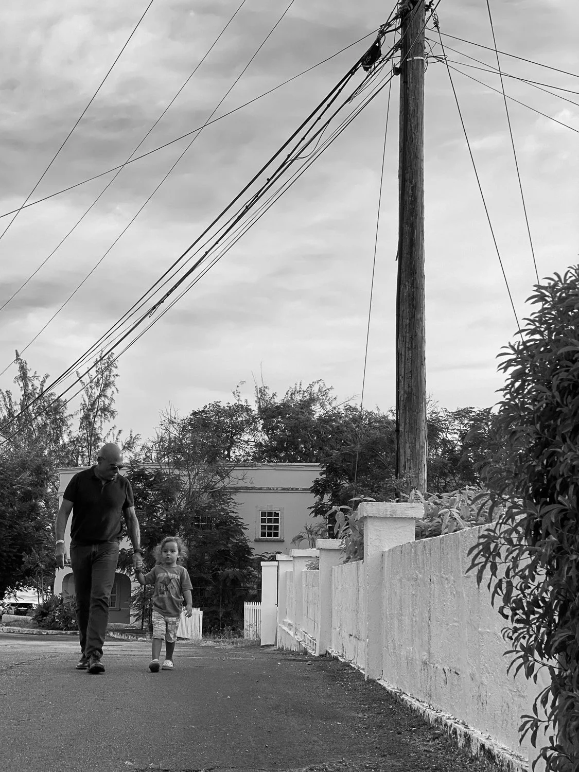 A man and a young girl walking along a residential street holding hands, with trees, a white fence, and utility pole with power lines in the background