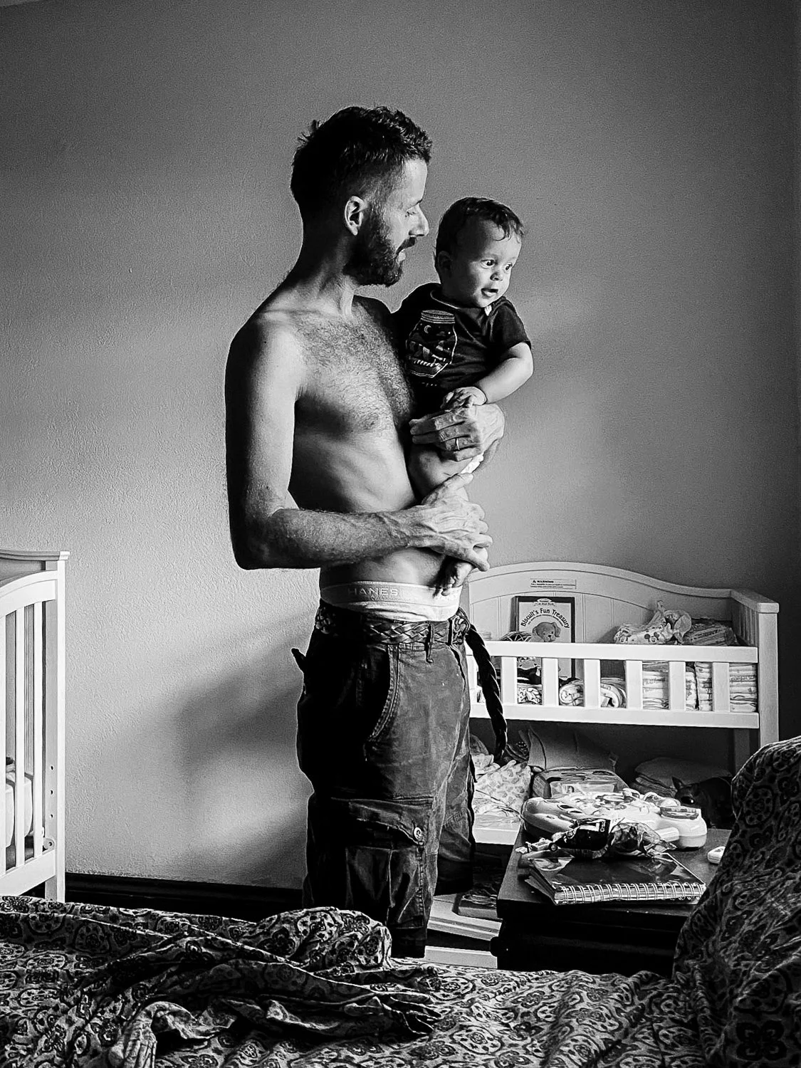 A shirtless man holding a young child in a toddler bed room.