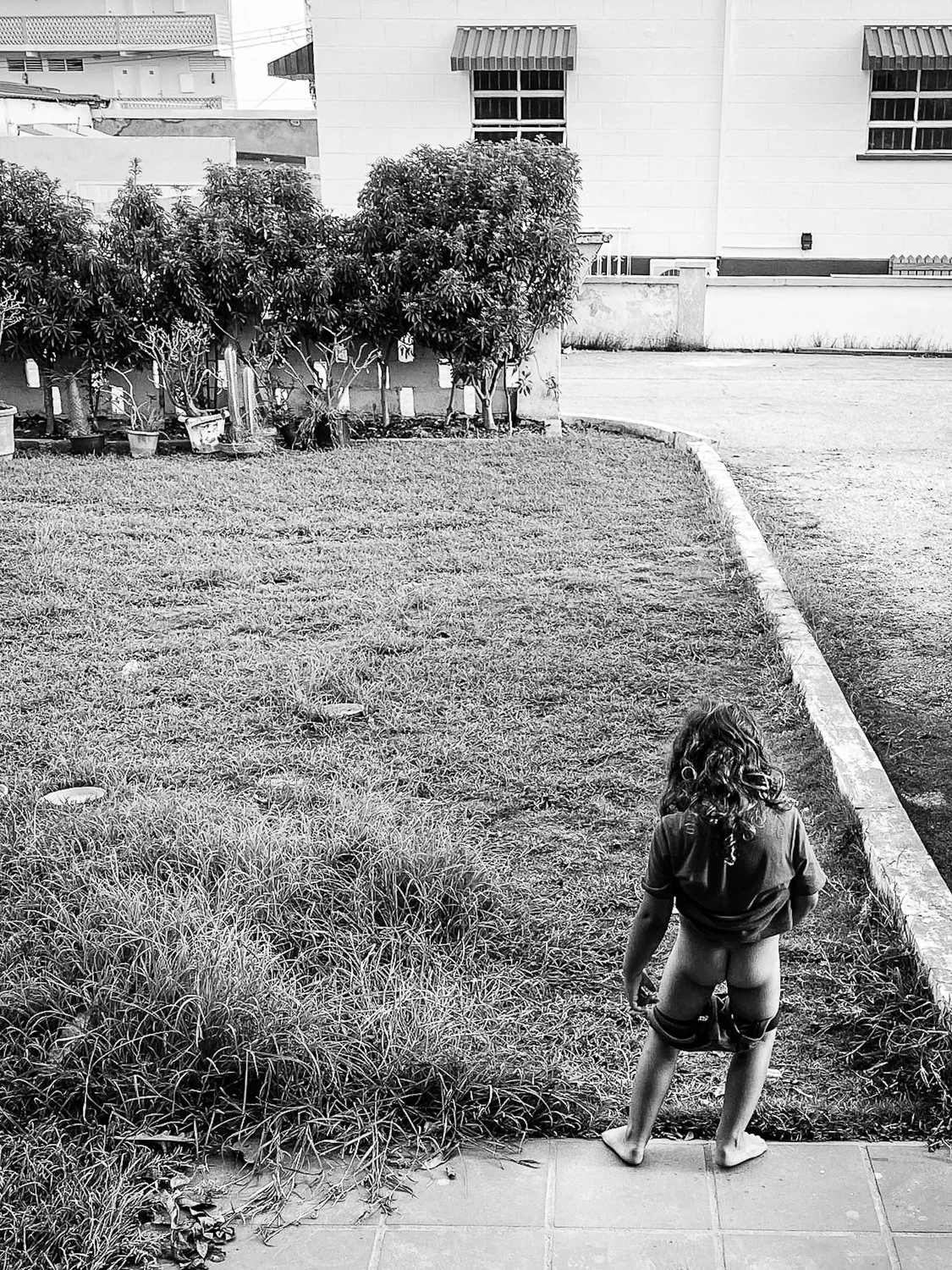 Child standing on a sidewalk, facing away from the camera, with shorts pulled down, in a backyard with grass, bushes, and houses in the background.