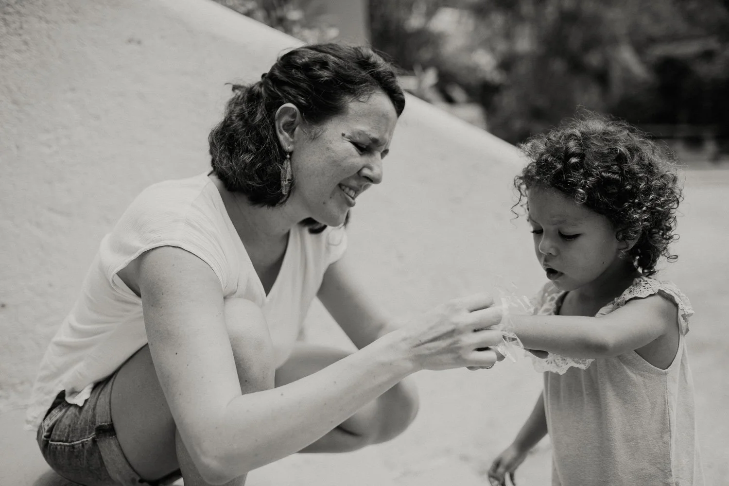 A woman and a young girl sharing a moment outside. The woman is smiling and engaged with the girl, gently touching the girl's arm as they look at each other. The image is in black and white.