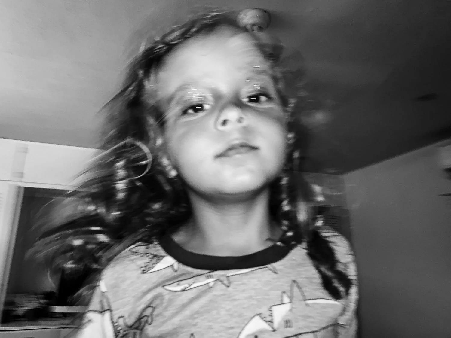 A young girl with curly hair is looking at the camera, with a slight smile, wearing a shirt with cartoon characters on it, in a room with blurred background.