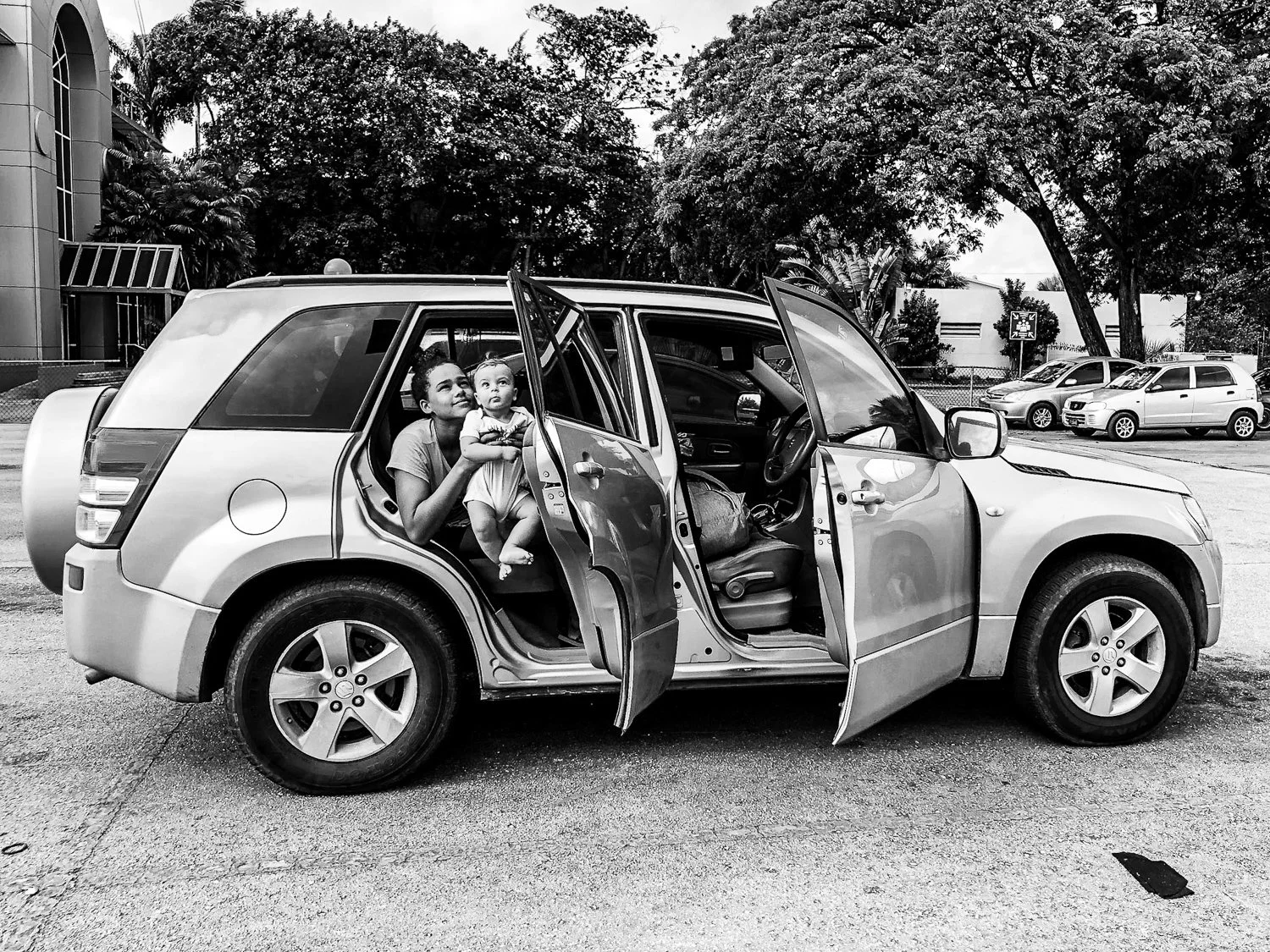 A woman and a young girl sitting in the open back seat of a parked SUV in a parking lot, with trees and other cars in the background.