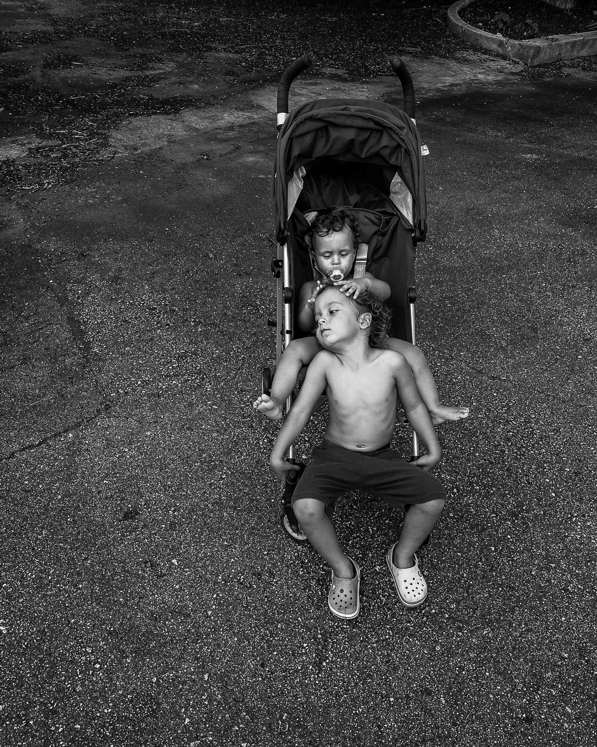 Two children resting in a stroller on a rough outdoor surface. One child is sitting upright with legs spread, wearing Crocs, and the other is lying with head on the first child's lap, with a pacifier in mouth and hand on his head.