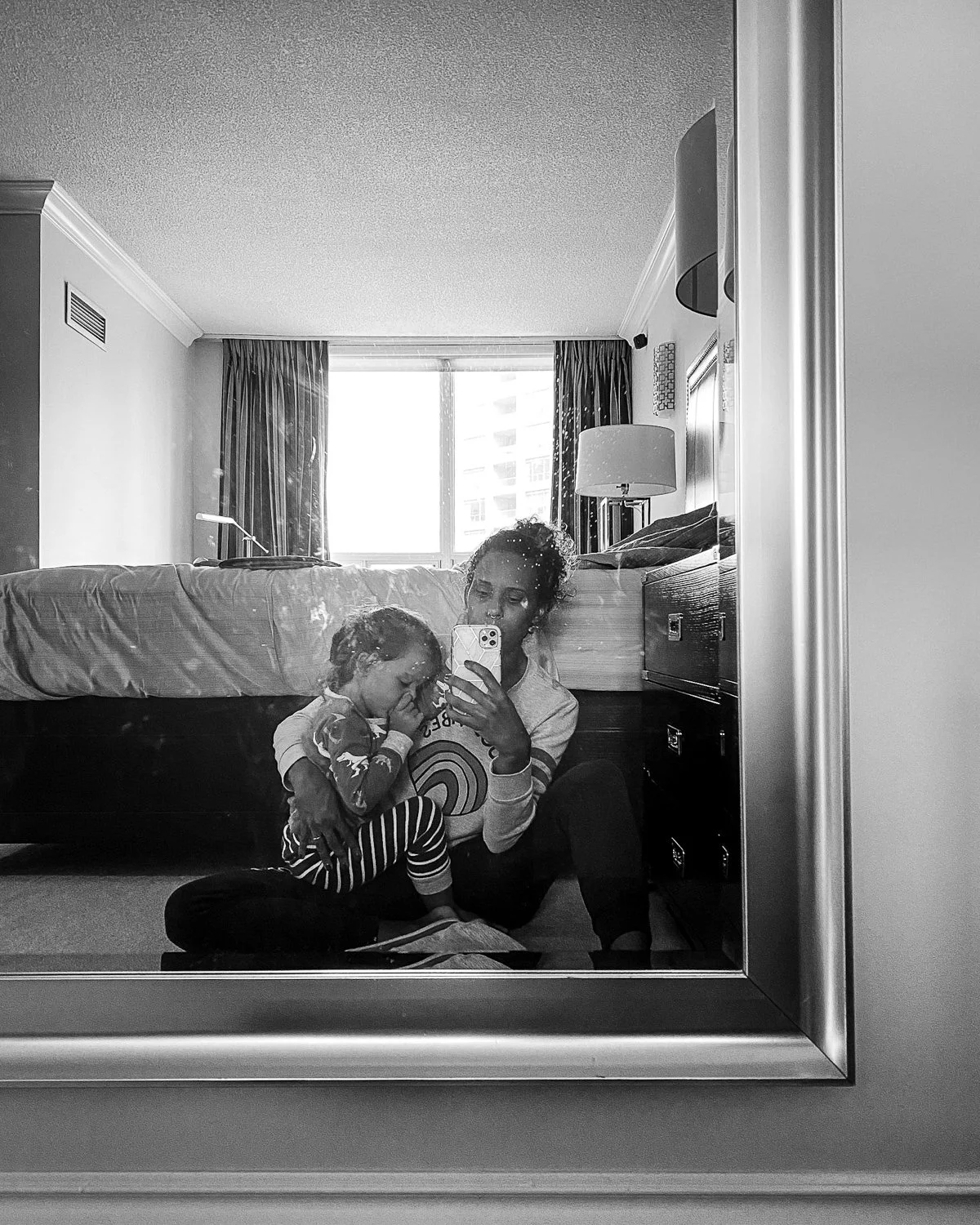 A woman and a young girl taking a selfie in a hotel room mirror. The woman is sitting on the floor, holding the phone, with her hair tied up. The girl is sitting on the woman's lap, snuggling with her and wearing striped pajamas.