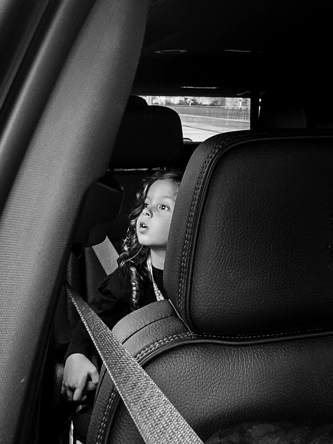 A young girl with curly hair looking out from the backseat of a car, captured from inside the vehicle with seats and seatbelt visible in black and white.