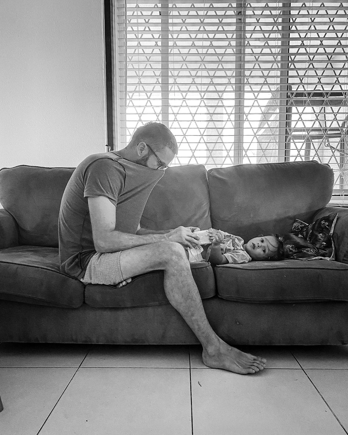 A man sitting on a couch, pulling his shirt over his mouth, looking at a young girl lying on the couch, looking back at him.