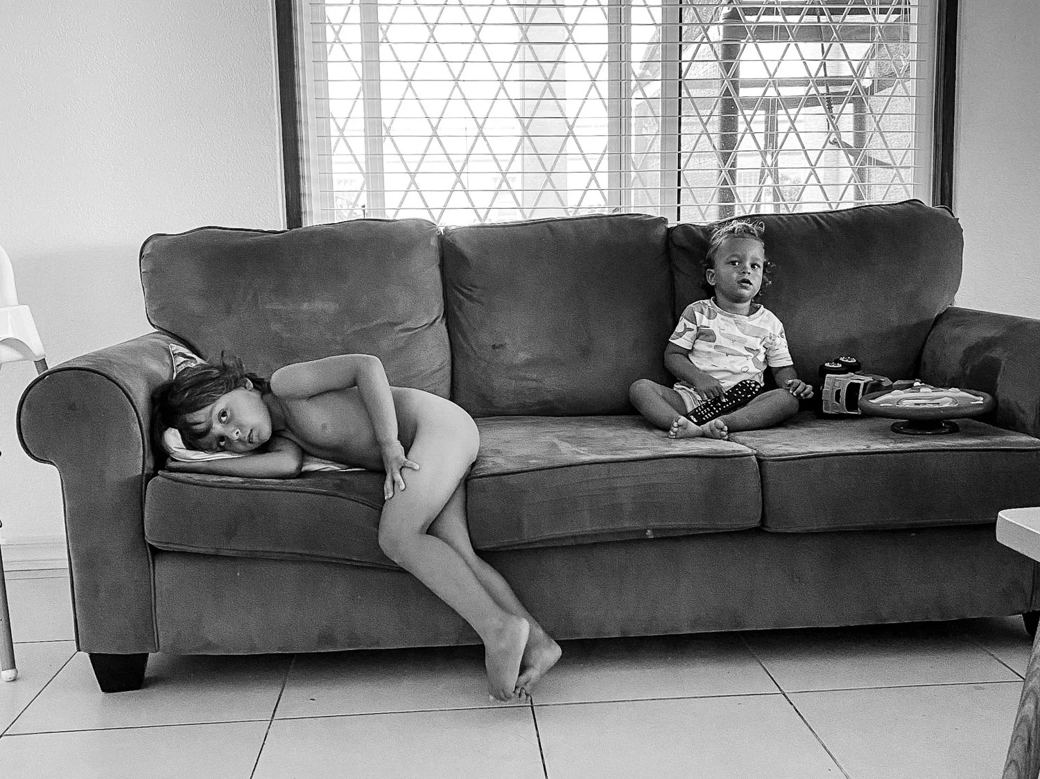 A black and white photo of two children on a couch in a living room. One child is lying on their side, looking at the camera, with long hair and no shirt. The other child is sitting upright, holding a remote control, wearing a patterned shirt and sho
