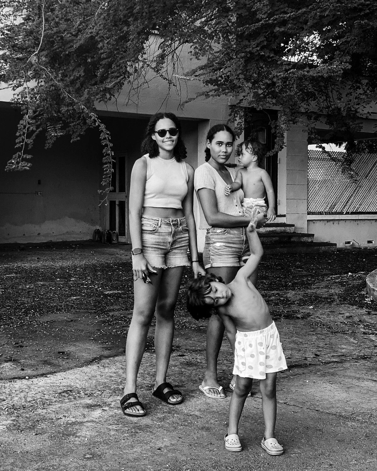 A black-and-white photo of three young women and two children standing outdoors in front of a house, with trees overhead. One woman is smiling and wearing sunglasses, while the other woman holds a child and gazes at the camera. A young boy stands in