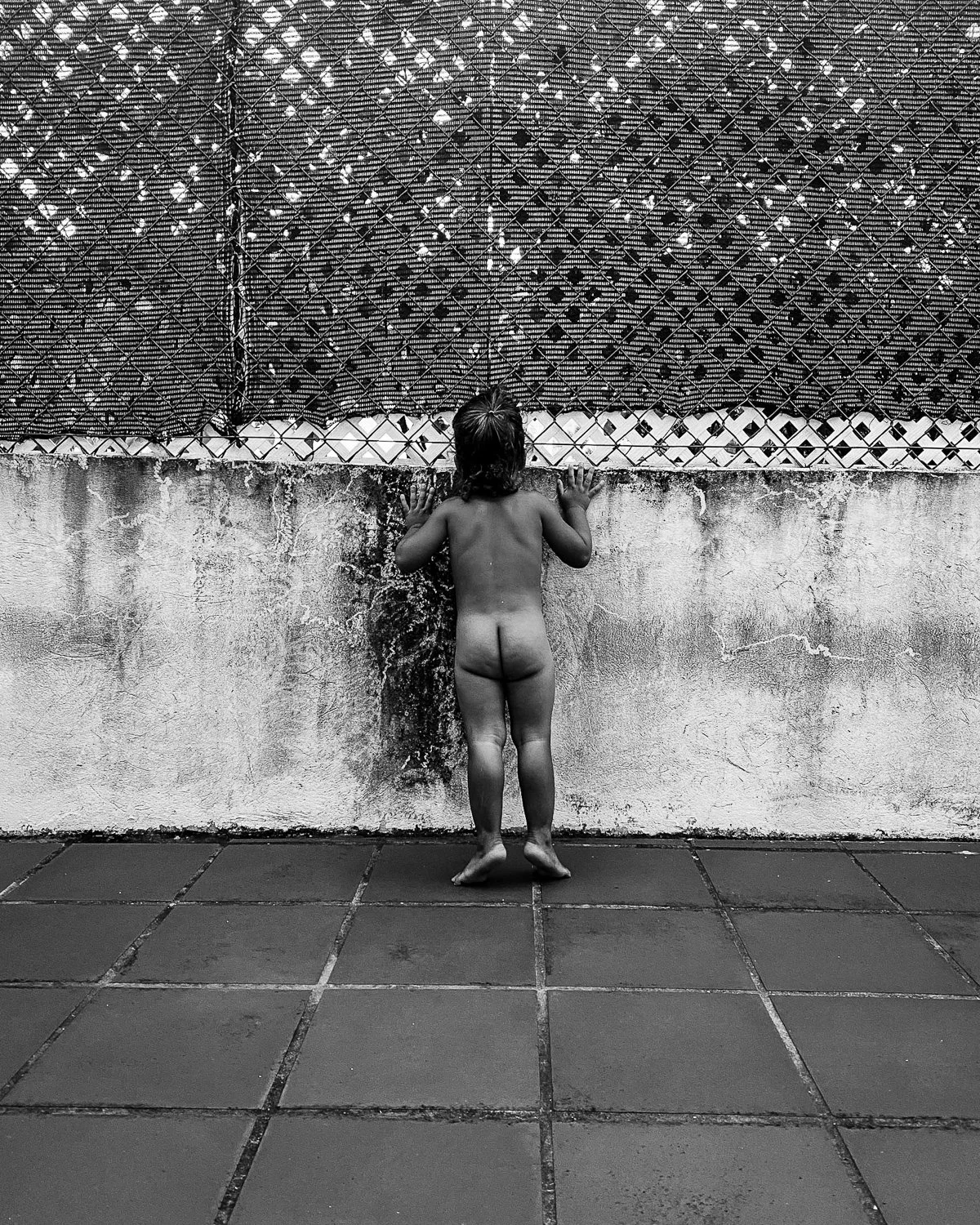 A young child standing naked on a tiled floor, facing a textured wall and a chain-link fence, with hands against the fence, water running down the child's back.