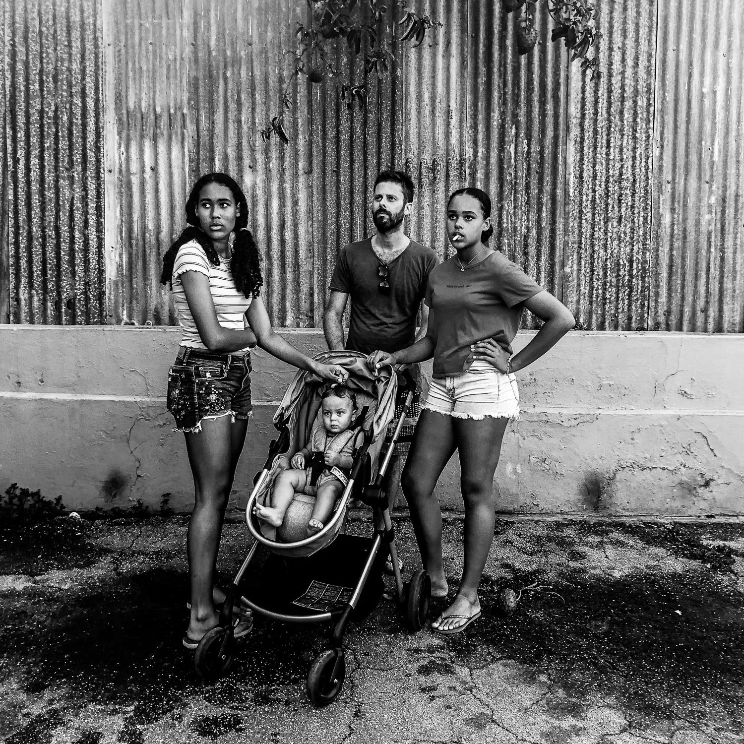 A black and white photo of three women and a man standing outdoors in front of a corrugated metal wall. One of the women is pushing a stroller with a young child sitting in it.