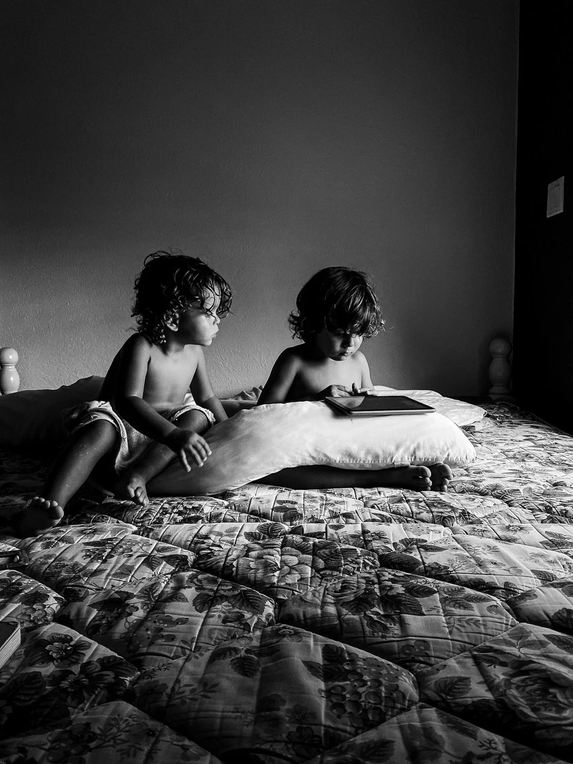 Two young children with curly hair sitting on a bed, one looking at a tablet and the other sitting on a pillow, in a bedroom with a plain wall.