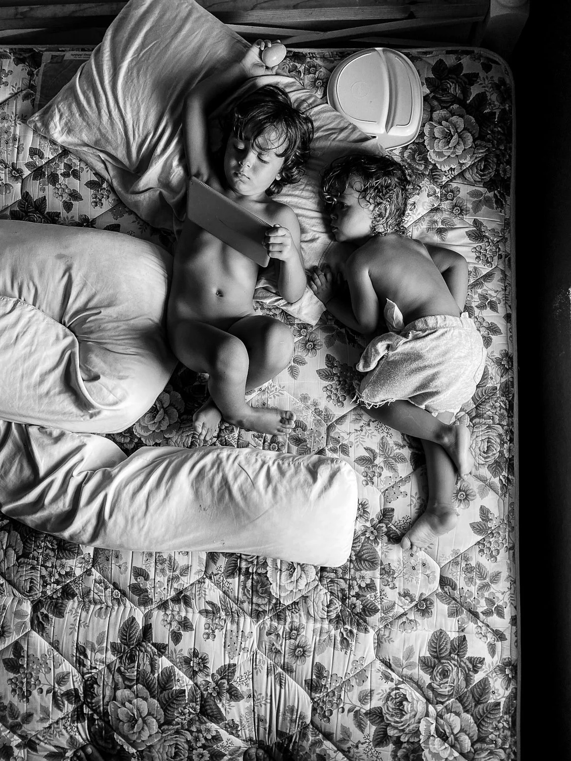 Two young children with curly hair are sleeping on a floral-patterned bed; one is using a tablet and holding a small object, while the other is curled up on their side.