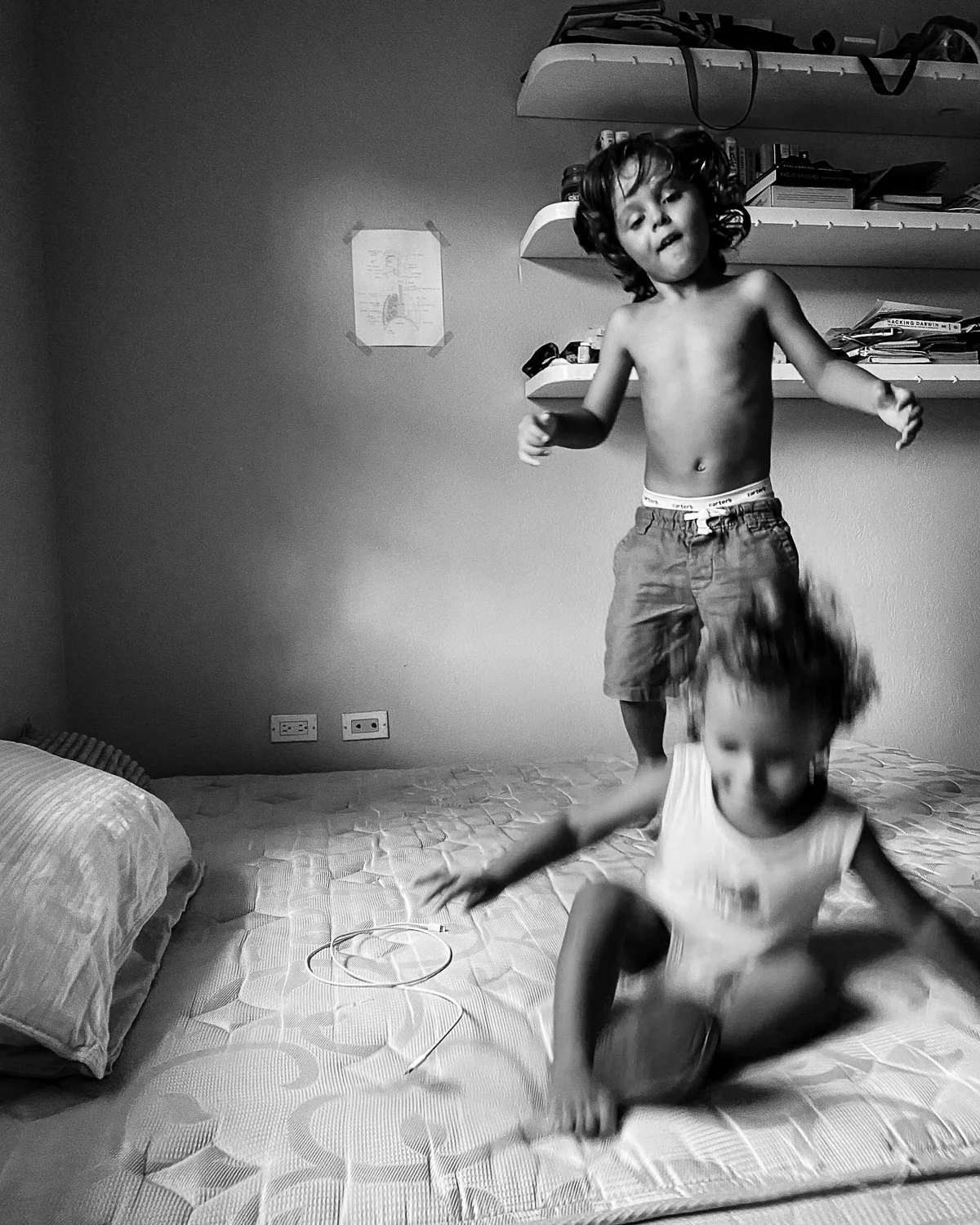 Two children playing on a bed in a bedroom. One child is jumping from the bed, and the other is sitting or squatting on the mattress. The room has shelves with books and items, a pillow, and electrical outlets on the wall.