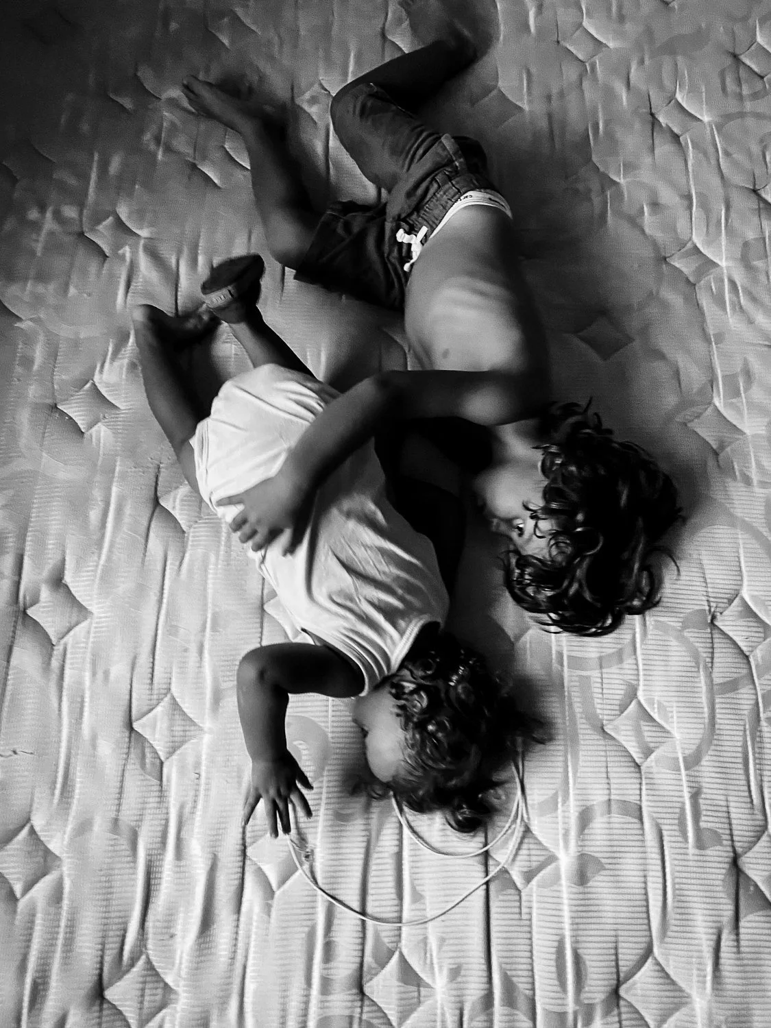 Two children lying on a mattress, embracing each other. One child is wearing a light shirt and shorts, the other in darker shorts. They are holding hands and facing each other.
