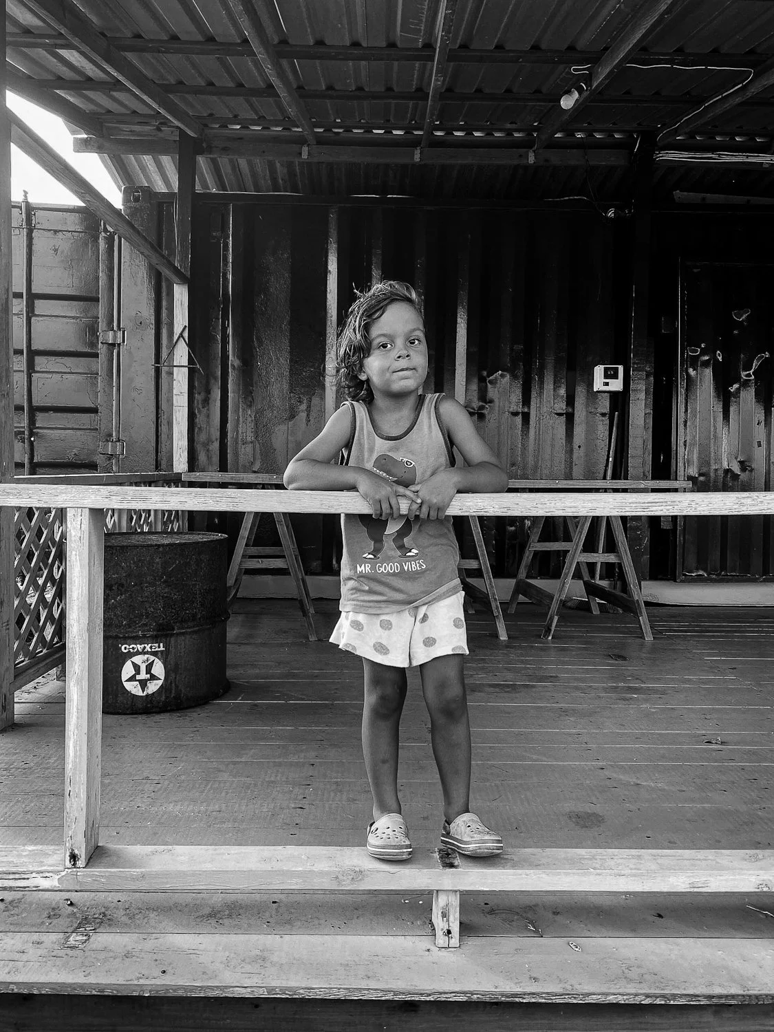 A young girl standing on a porch railing, looking at the camera with a serious expression. She is wearing a tank top with a dinosaur graphic and the words "Mr. Good Vibes," shorts with polka dots, and sneakers. The porch has wooden flooring and a rus