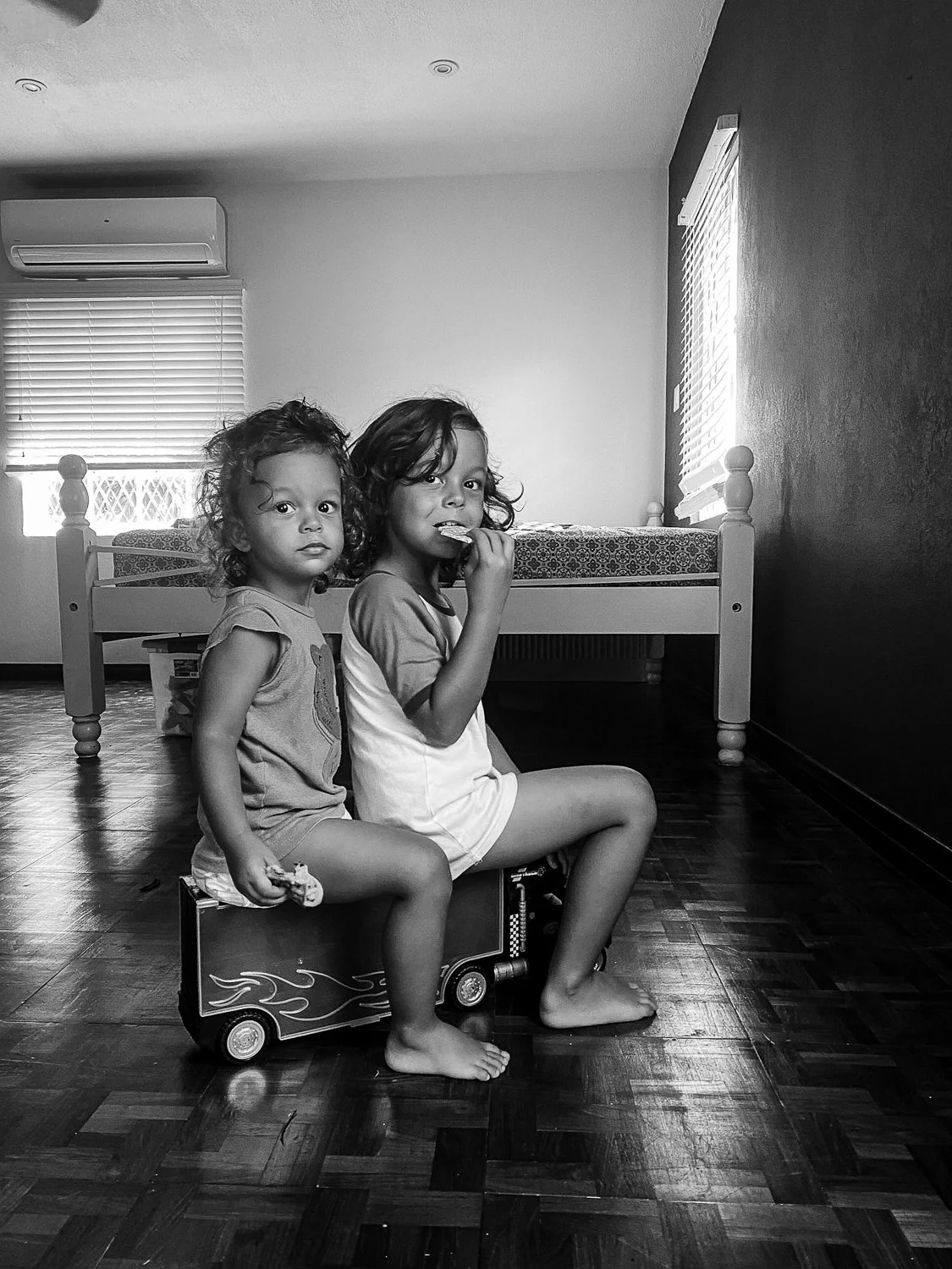 Two young girls sitting on a toy truck in a bedroom with hardwood floors, with one girl eating a snack and the other holding a toy, and a bed and windows with blinds in the background.