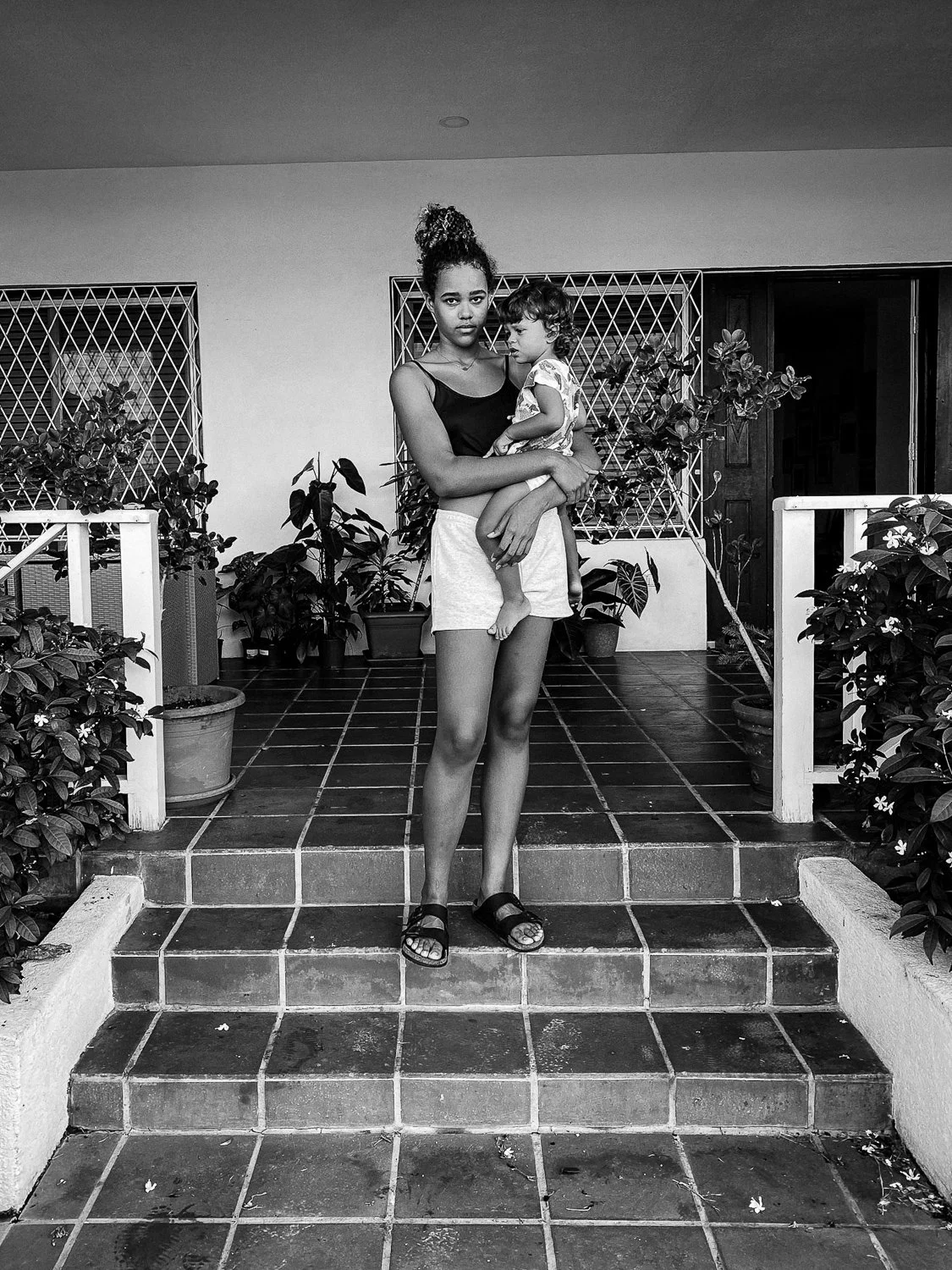 A woman holding a young girl on her lap standing on front steps of a porch with potted plants, lattice windows, and a door in the background.