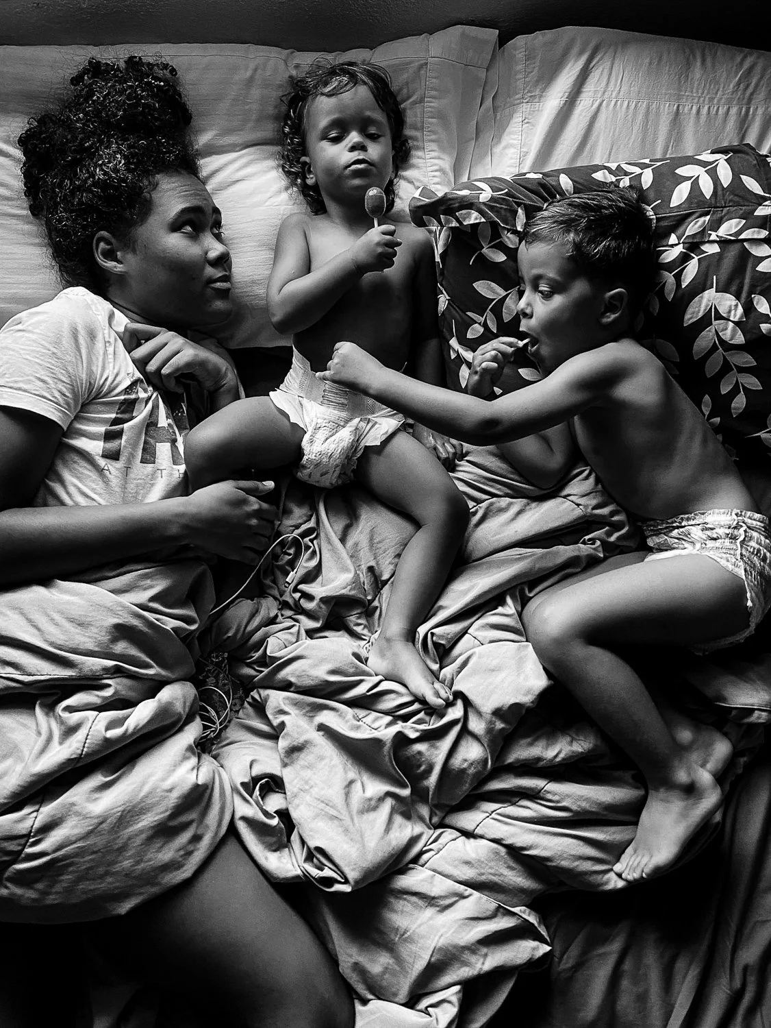 A woman and two young boys lying on a bed, with the boys playing and one holding a lollipop, in a black and white photograph.