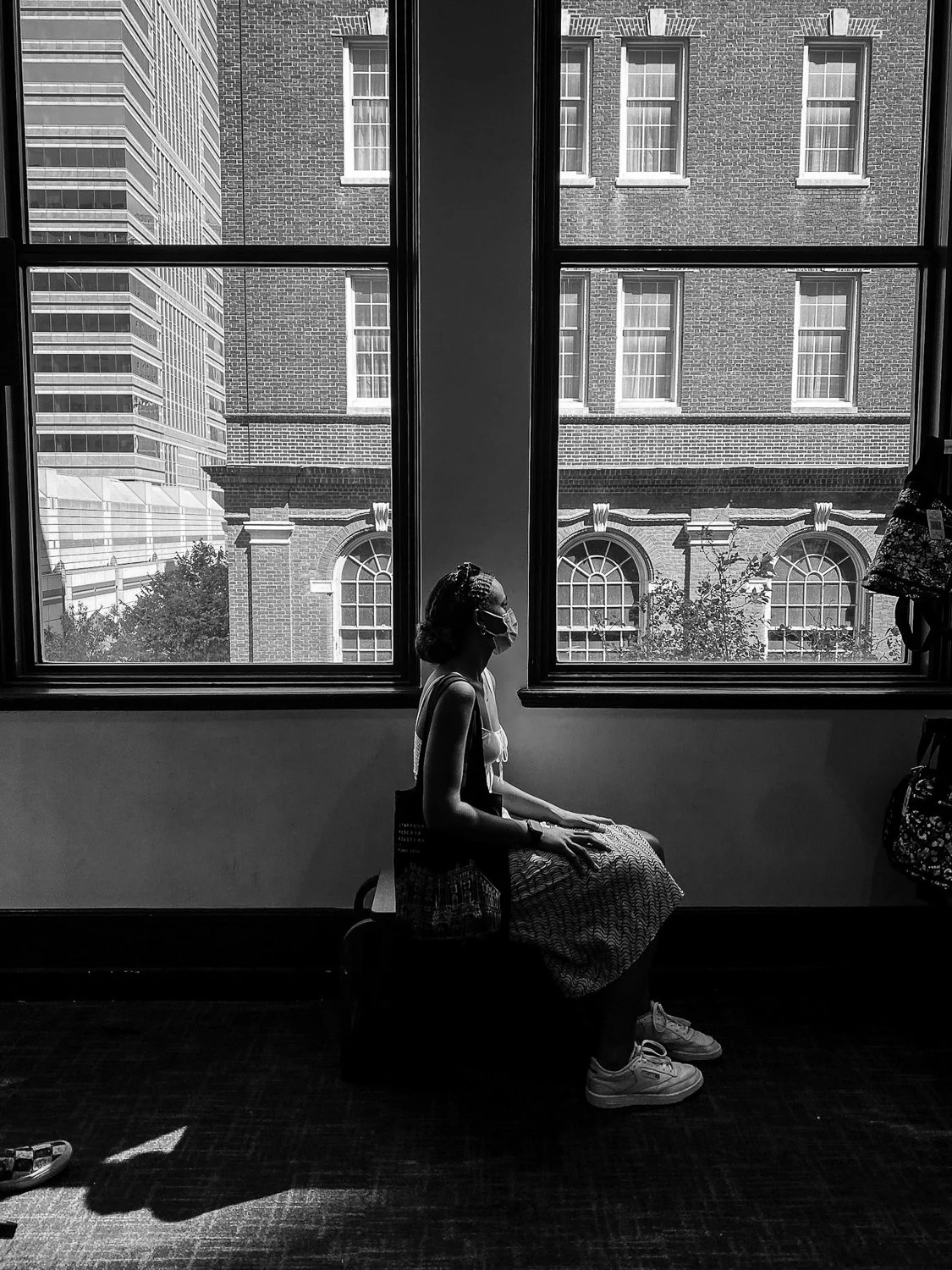 A woman sitting indoors on a box, wearing a face mask, with large windows behind her revealing a city scene with brick and glass buildings and trees outside.