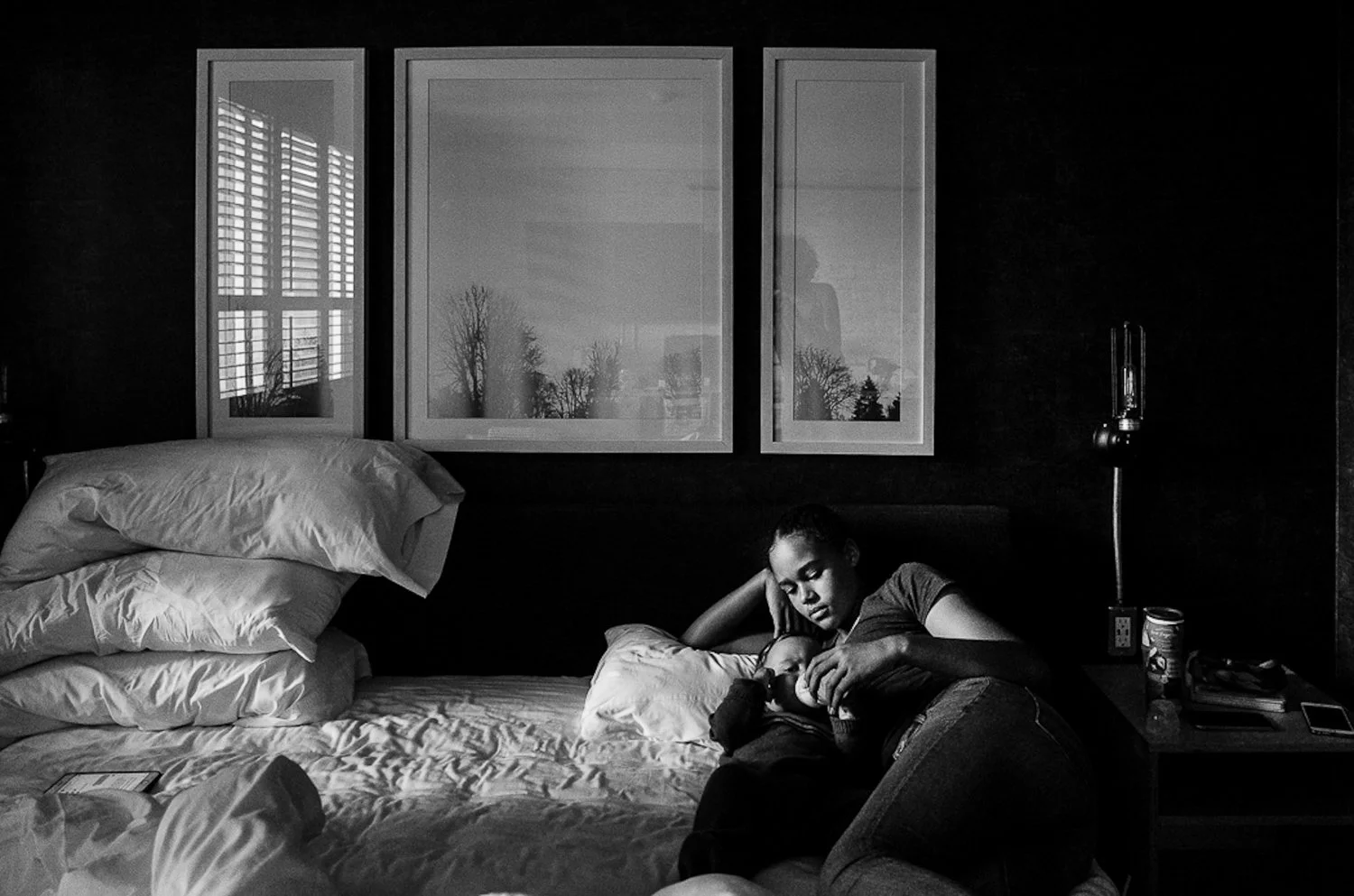 A woman lying on a bed holding a baby, in a bedroom with three picture frames on the wall and a nightstand with a lamp, a bottle, and a phone.