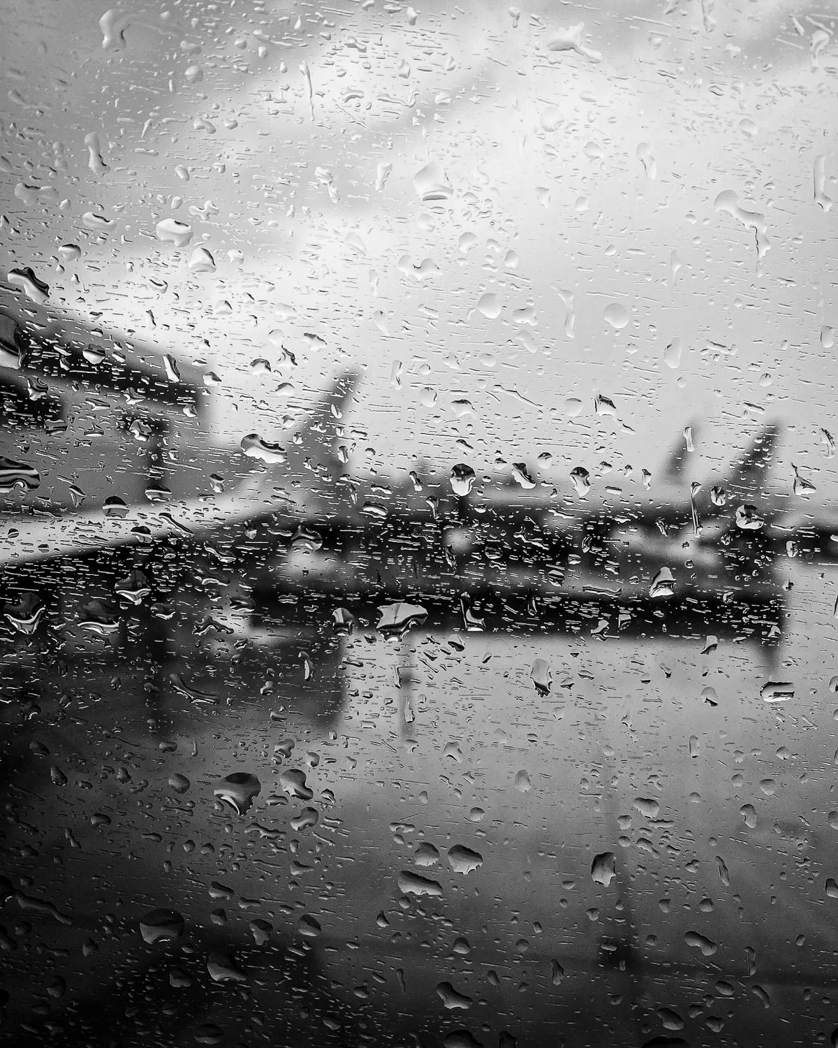 View of airplanes parked on a wet tarmac through a window with raindrops on the glass.