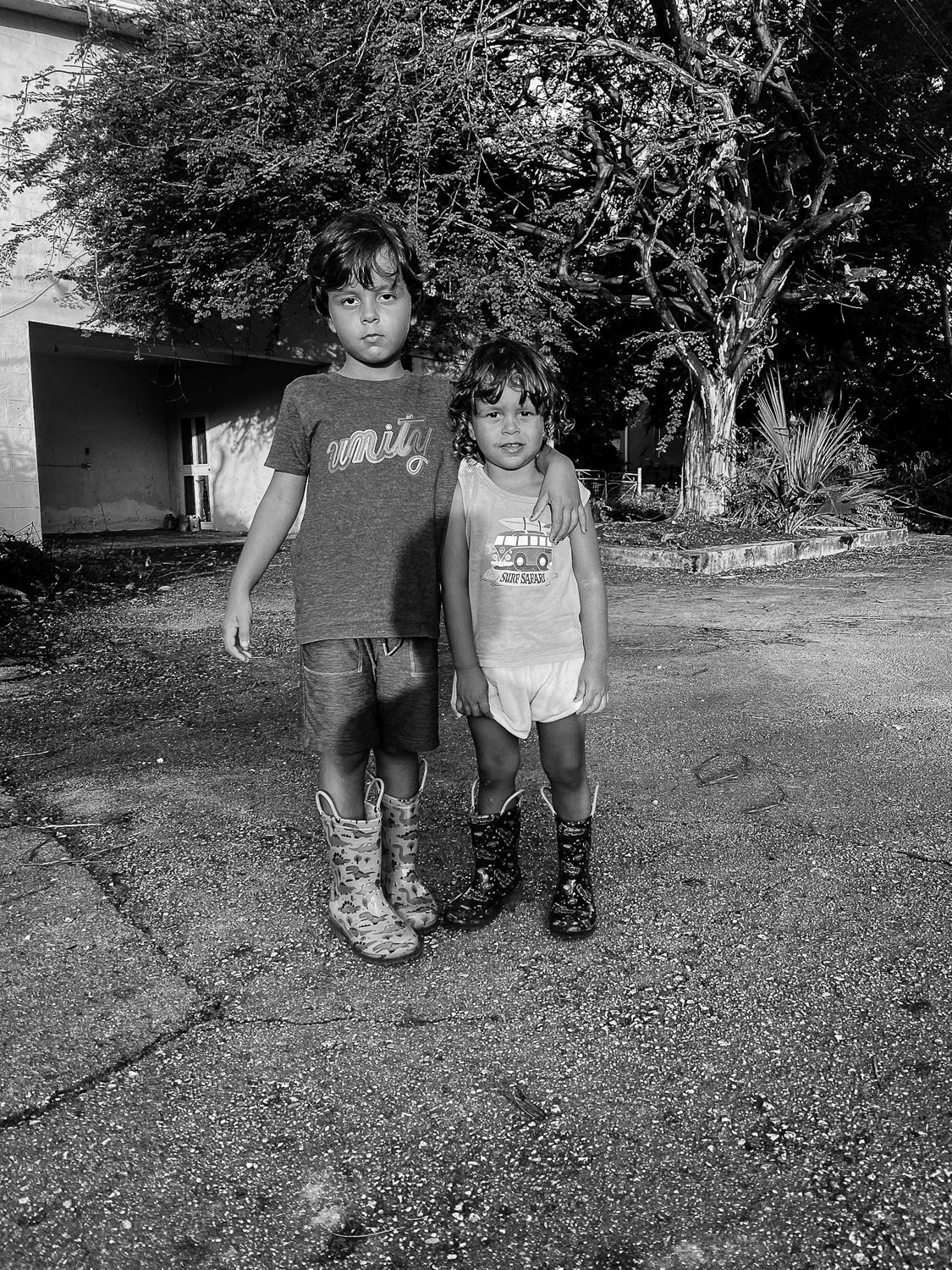 Two children standing outdoors on a rough, paved ground with a large tree and house in the background. The boy on the left is taller and wearing camouflage rain boots, a T-shirt, and shorts. The girl on the right is shorter, wearing patterned rain bo