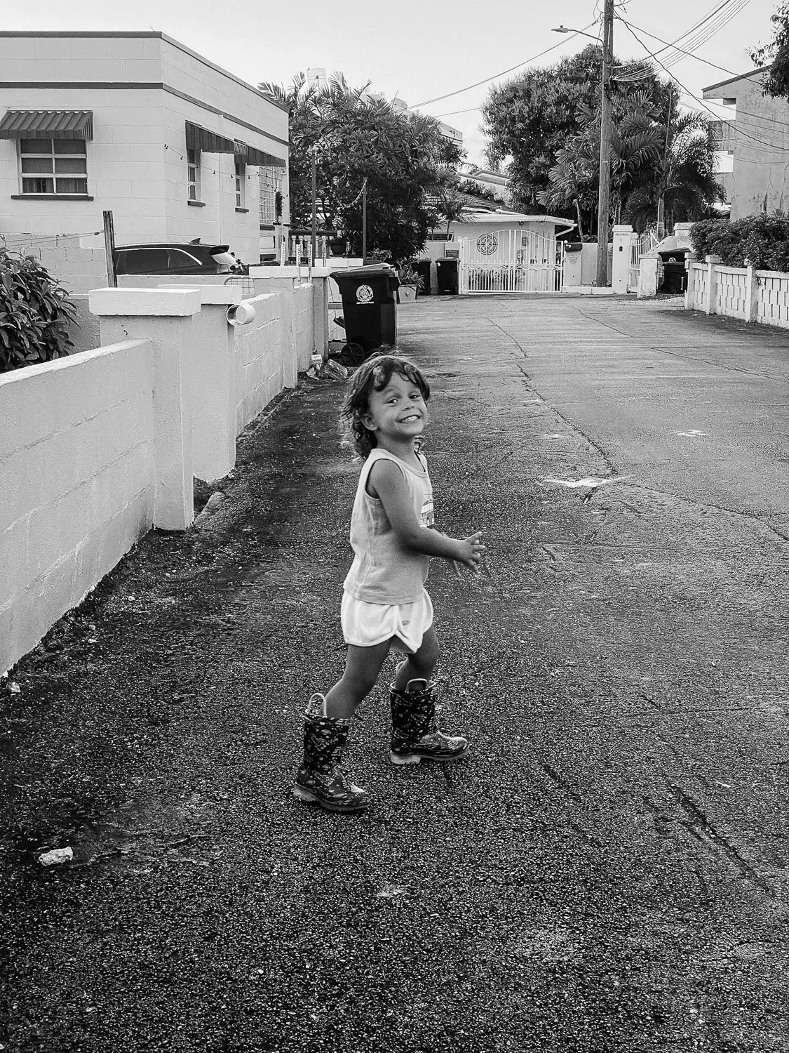 A young girl smiling and running on a wet sidewalk in a residential neighborhood, wearing rain boots and a sleeveless top and shorts, with houses, trees, and utility poles in the background.