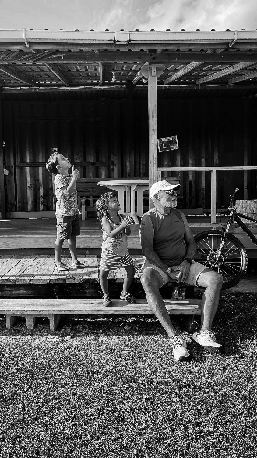 An older man with a beard, sunglasses, and a hat sitting on a wooden deck with two young children, one girl standing on the deck and one boy standing below it. Two bicycles are nearby, and the scene appears to be outdoors with a grassy area in the fo