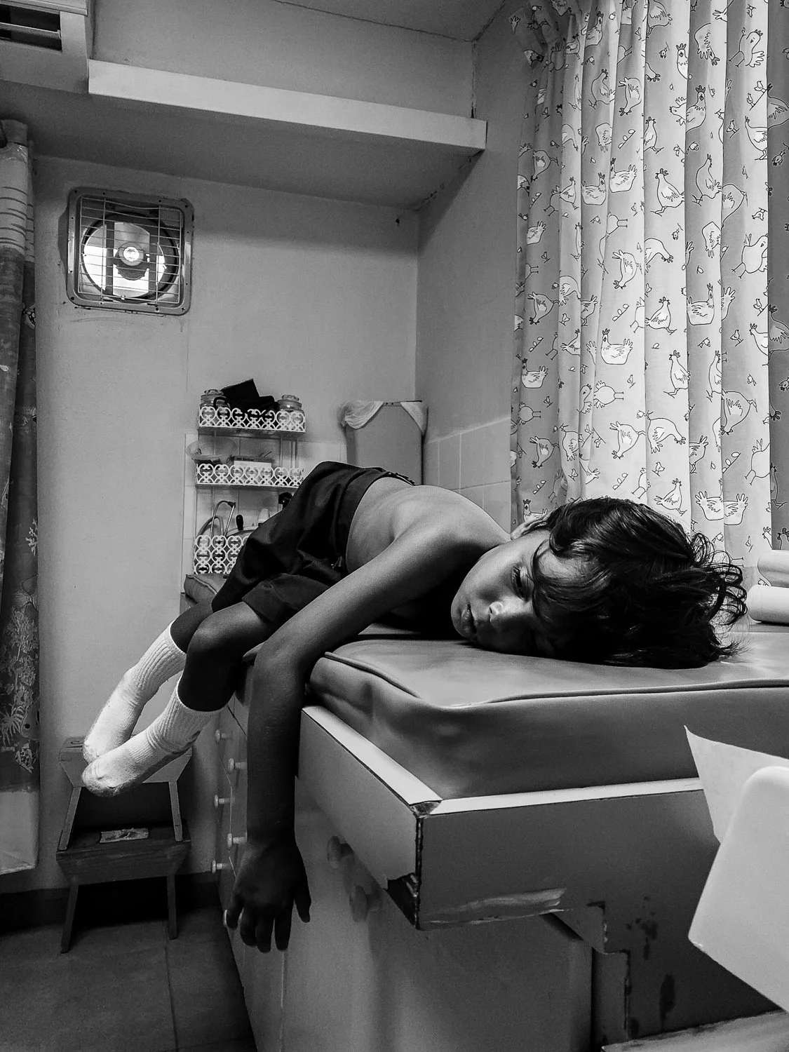 A young person lying face down on a bed in a small room, wearing socks and shorts, with dark hair and looking towards the camera.